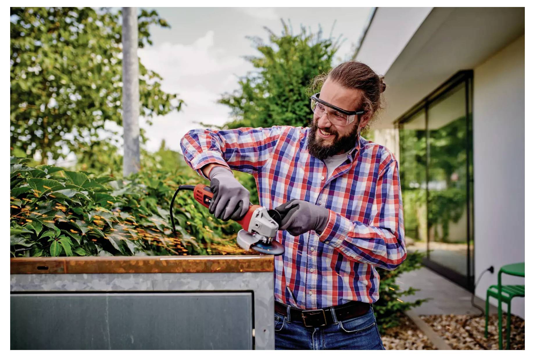 Mann mit Brille und Handschuhen, der eine Hecke mit einem elektrischen Werkzeug beschneidet und draußen in einer Gartenumgebung neben einem modernen Gebäude steht.