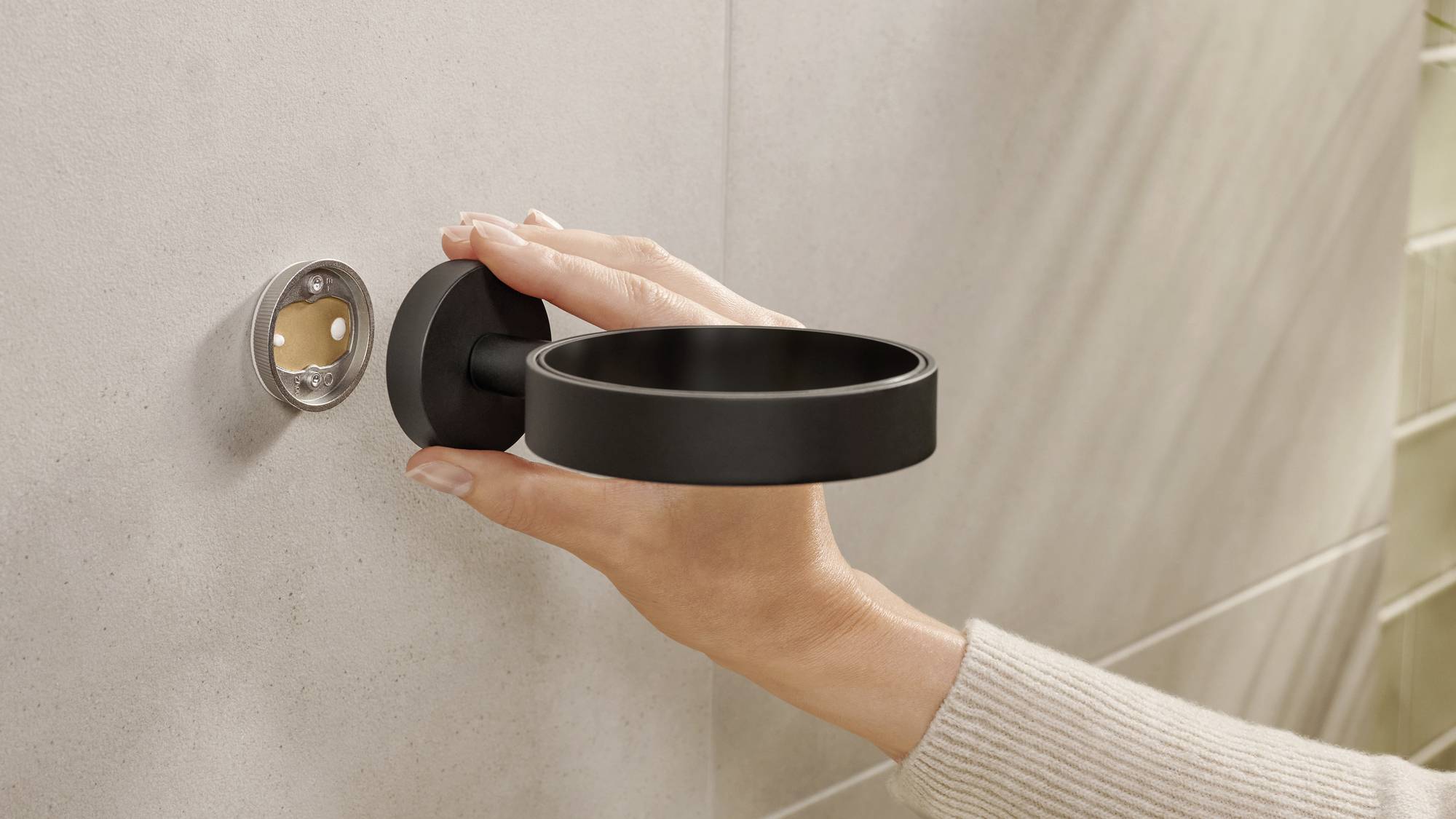 Eine Hand montiert einen schwarzen Halter an einer grauen Wand, vermutlich für einen Badezimmer-Accessoire wie einen Becher oder Seifenspender.