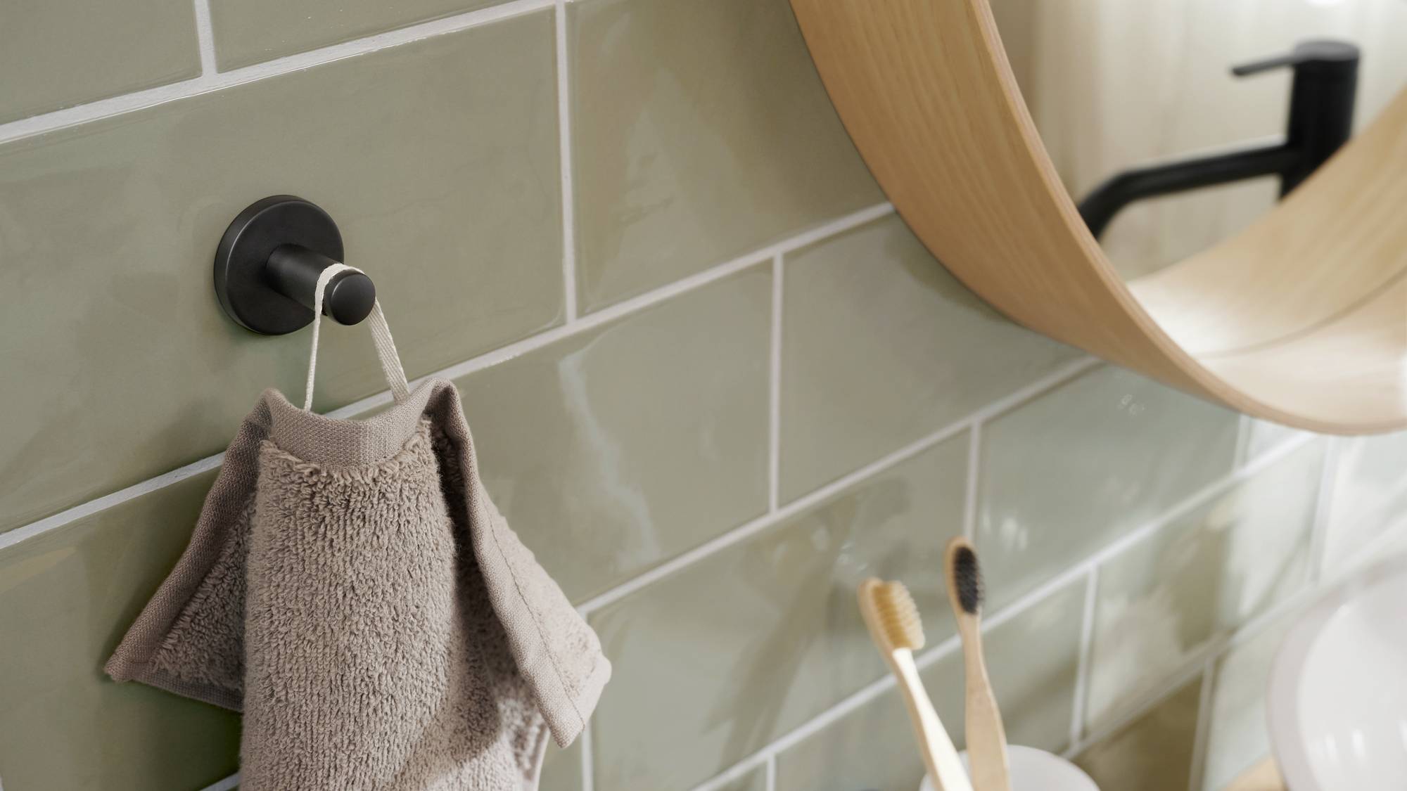 Handtuch hängt an einem Haken an einer gefliesten Badezimmerwand, daneben ein runder Spiegel und zwei Zahnbürsten im Halter auf dem Waschbecken.