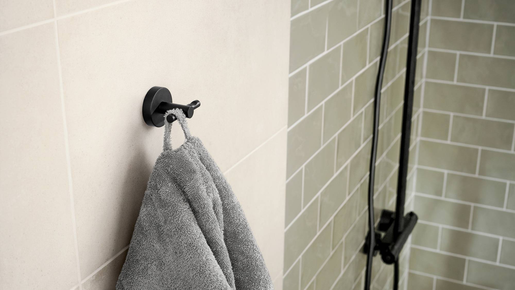 Ein graues Handtuch hängt an einem runden, schwarzen Wandhaken im Badezimmer mit Fliesenwänden in beigen und graugrünen Tönen.