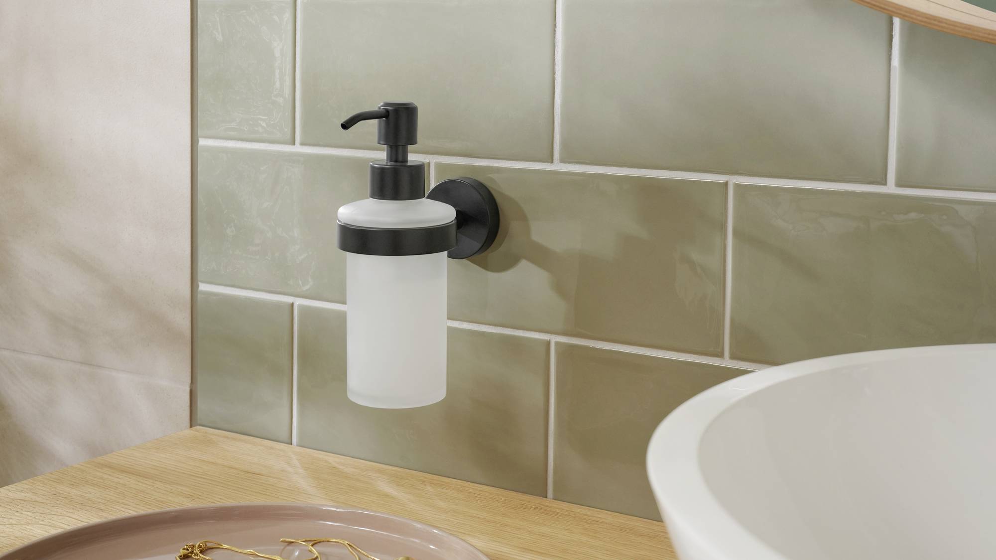 Ein Seifenspender ist an einer Wand mit beigefarbenen Fliesen neben einem Waschbecken angebracht. Im Vordergrund liegt ein Tablett mit Schmuck.