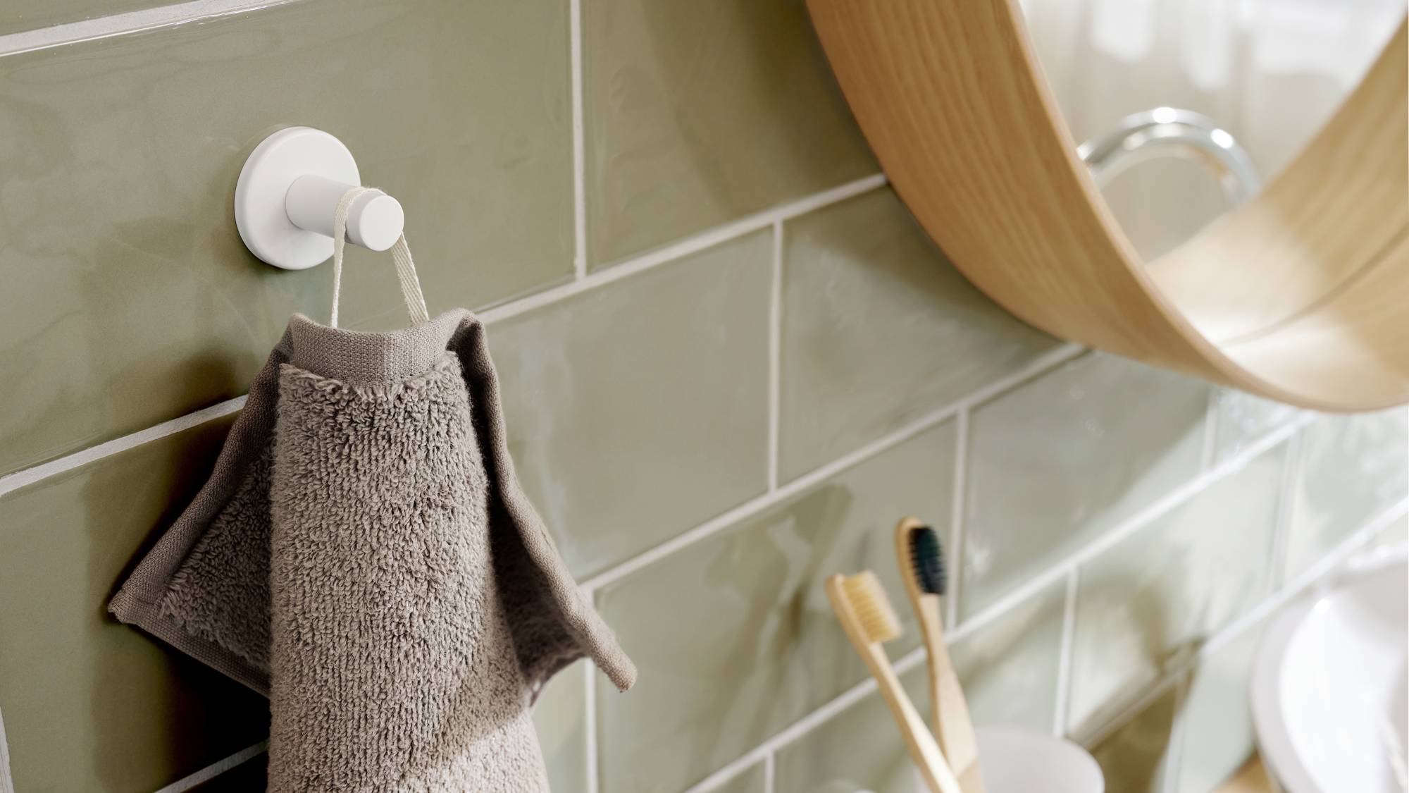 Braunes Handtuch hängt an einem weißen Haken auf grünen Fliesen. Im Hintergrund ein Spiegel und zwei Holzzahnbürsten im Zahnputzbecher.