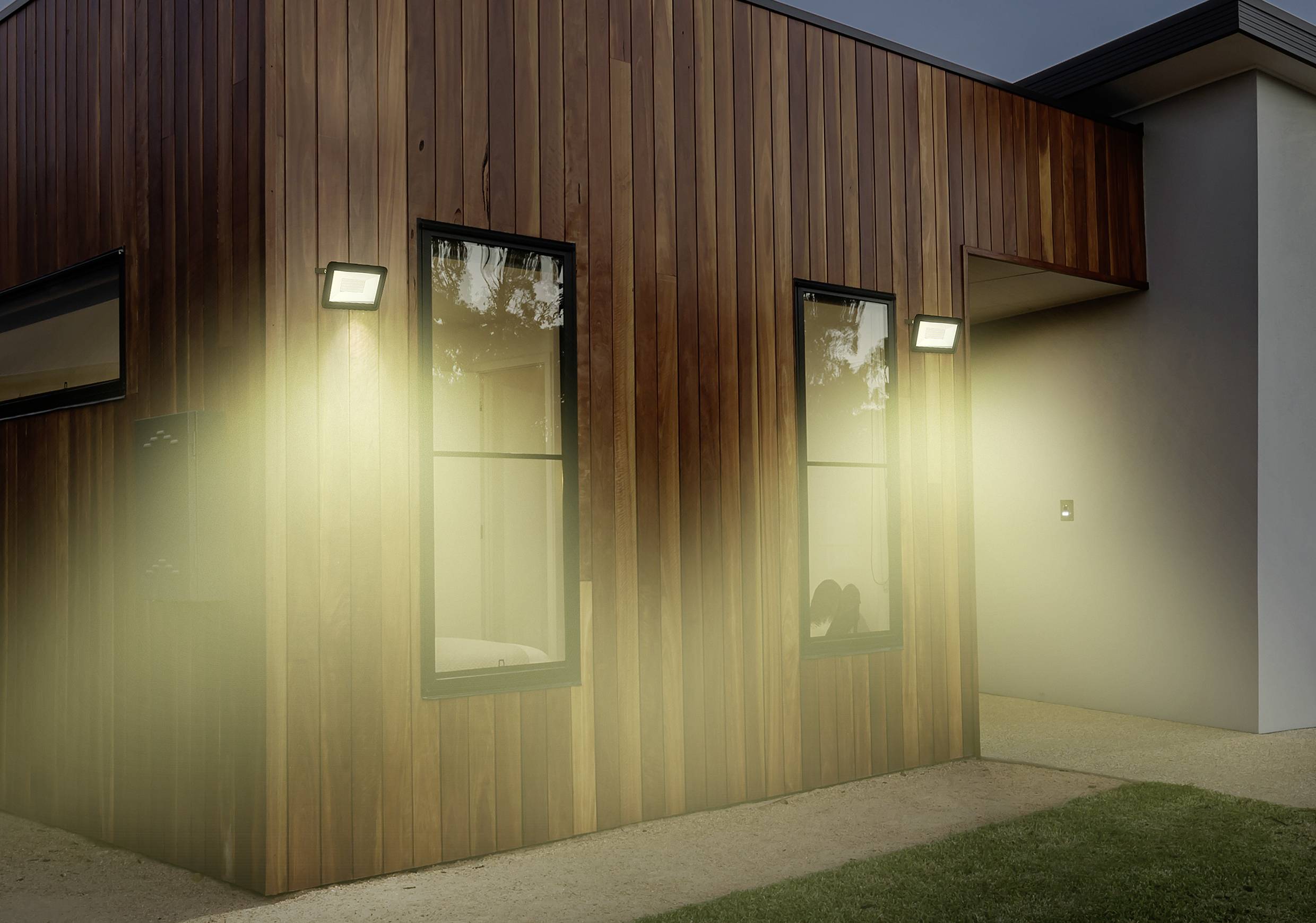 Ein modernes Holzhaus mit eingeschalteten Außenleuchten bei Dämmerung. Schlichte Architektur mit klaren Linien und Fensterfronten.