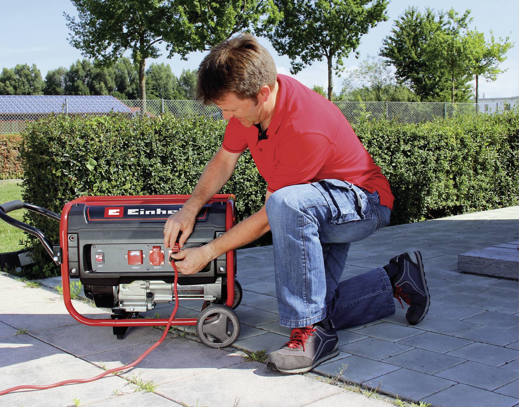 Ein Mann kniet und bedient ein rot-schwarzes Elektroaggregat im Freien, umgeben von Pflanzen und einem gepflasterten Weg.