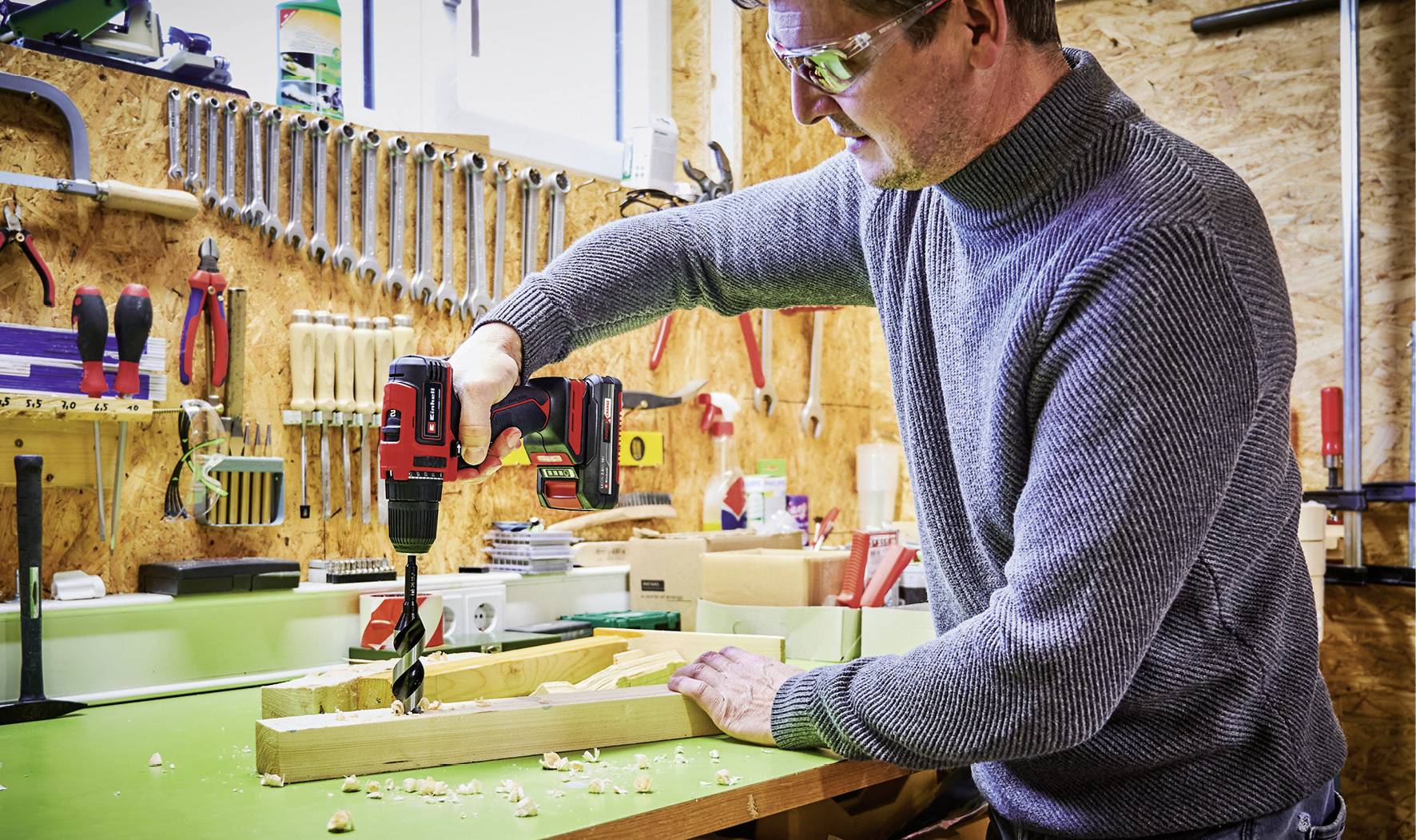 Mann in Werkstatt bohrt mit Akkubohrer ein Stück Holz. Hintergrund zeigt Werkzeuge an der Wand, darunter Schraubenschlüssel und Hämmer.