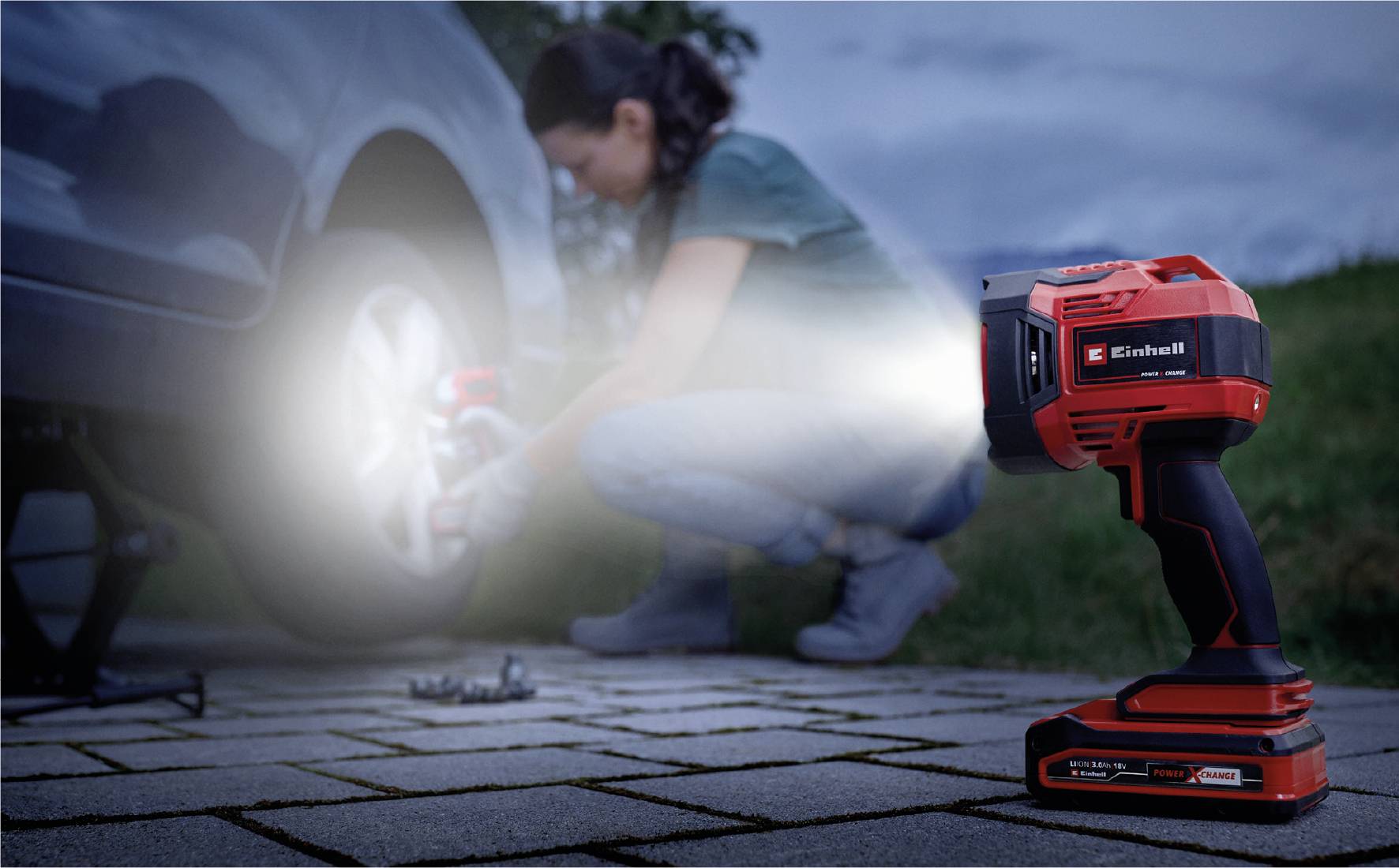 Eine Person wechselt am Abend ein Autoreifen mit Hilfe einer Taschenlampe, die das Rad ausleuchtet. Im Hintergrund ist leichtes Gras zu sehen.