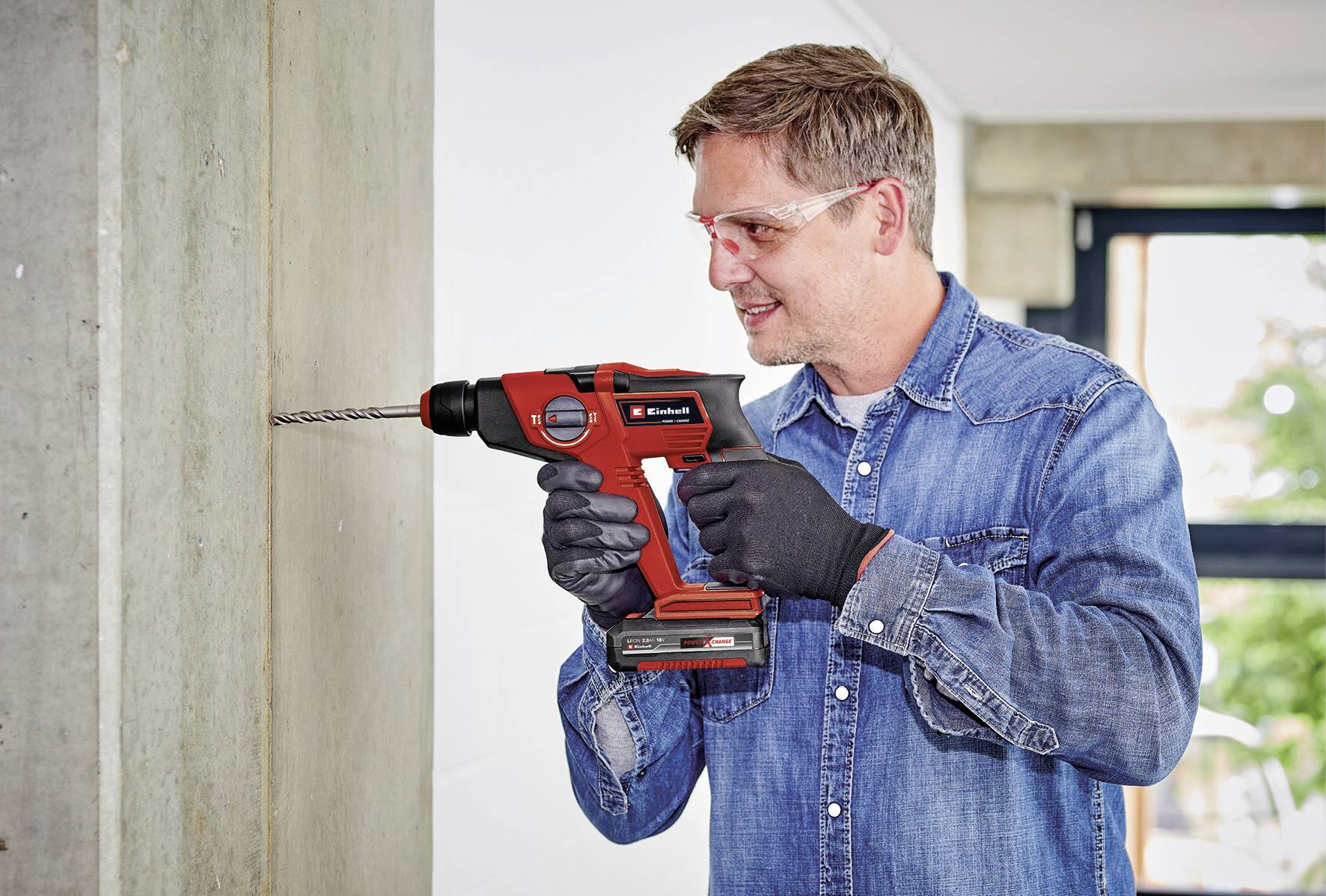 Ein Mann bohrt mit einer roten Bohrmaschine in eine Betonwand. Er trägt Schutzbrille und Handschuhe, ist in Jeans gekleidet.