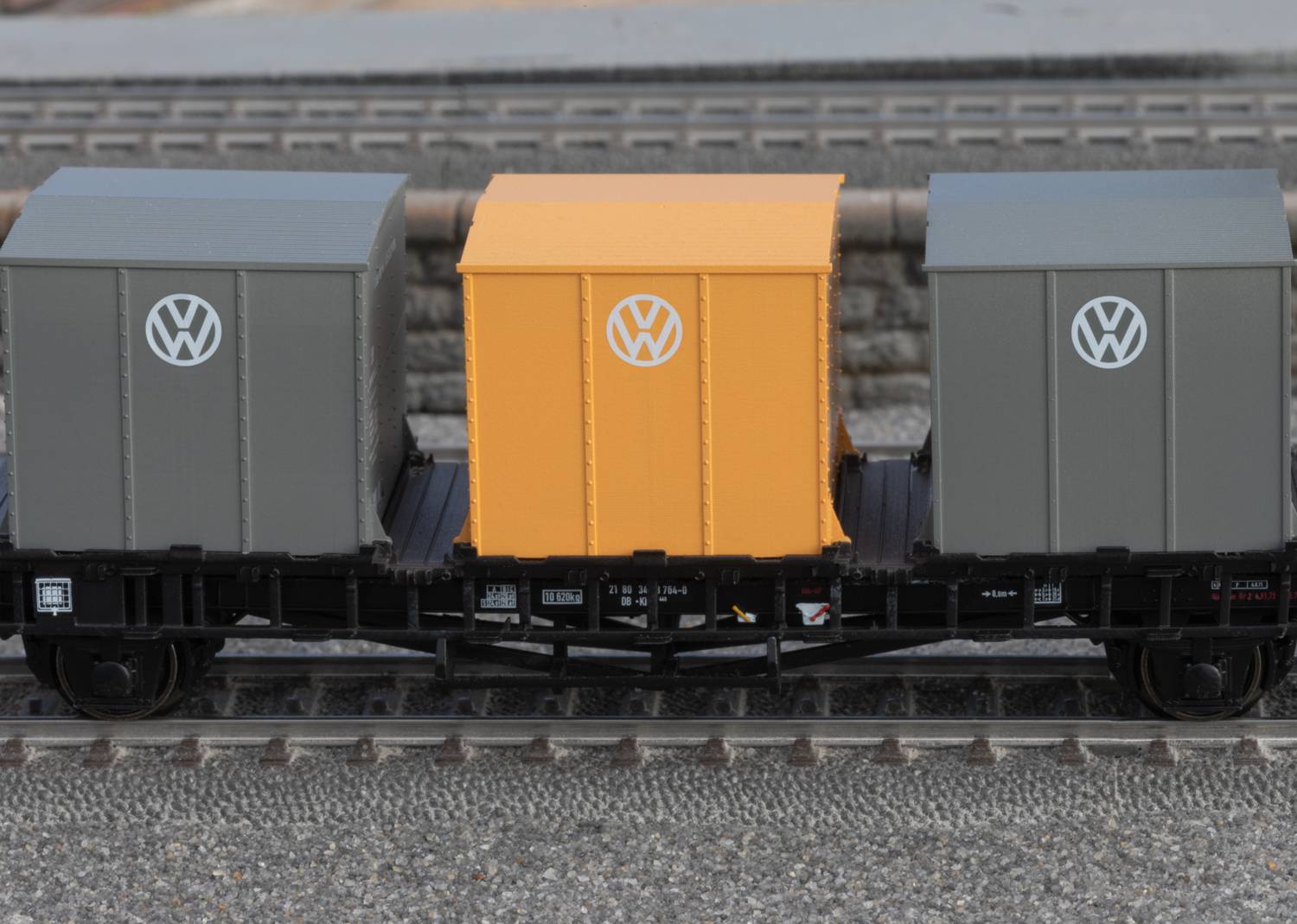Ein Modellzug mit drei Frachtcontainern, zwei graue mit VW-Logo und ein gelber in der Mitte, fährt auf einer Modelleisenbahnstrecke.