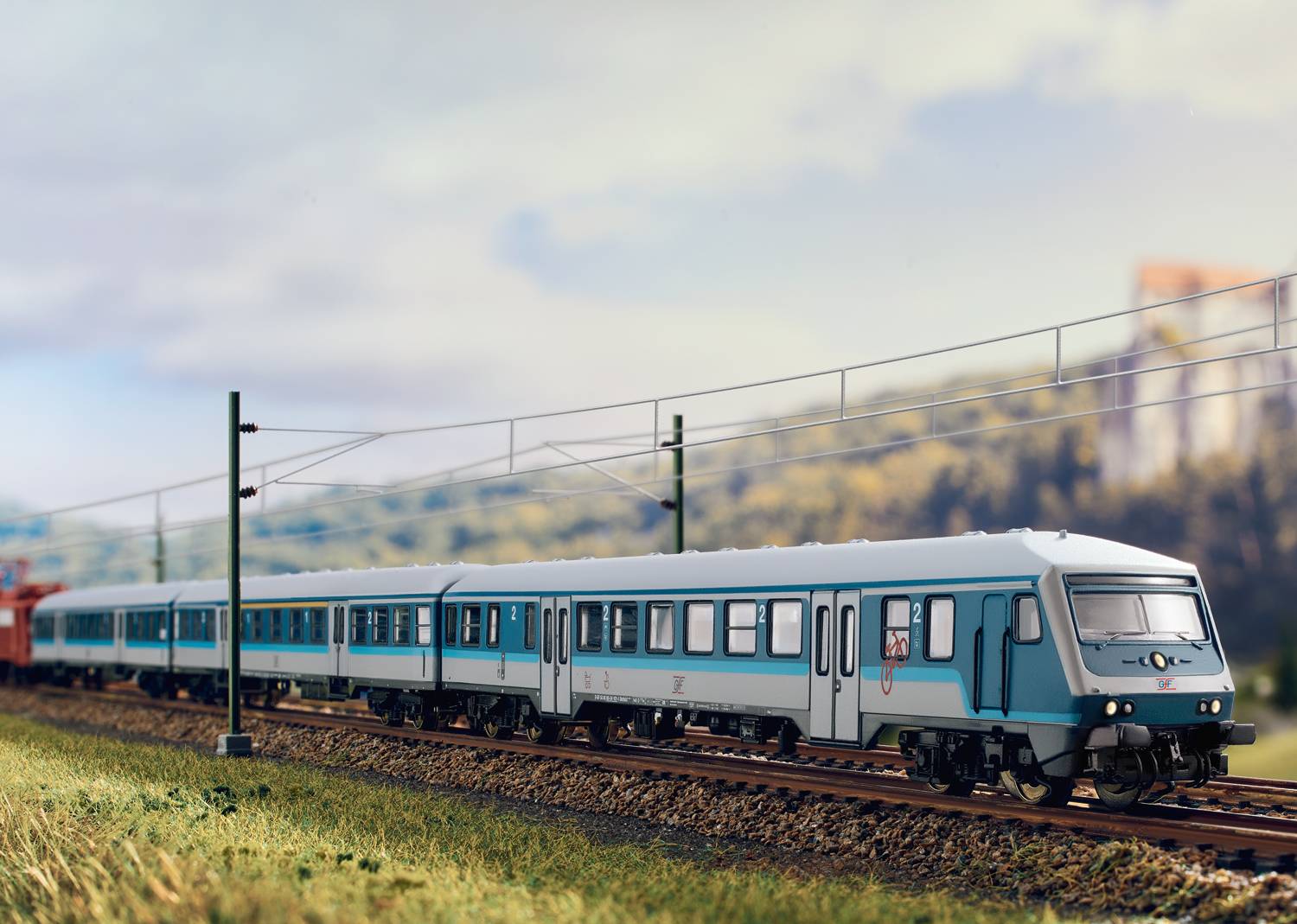 Ein blauer Zug fährt über Gleise auf einer Strecke durch eine ländliche Landschaft. Stromleitungen verlaufen parallel zur Bahnlinie.