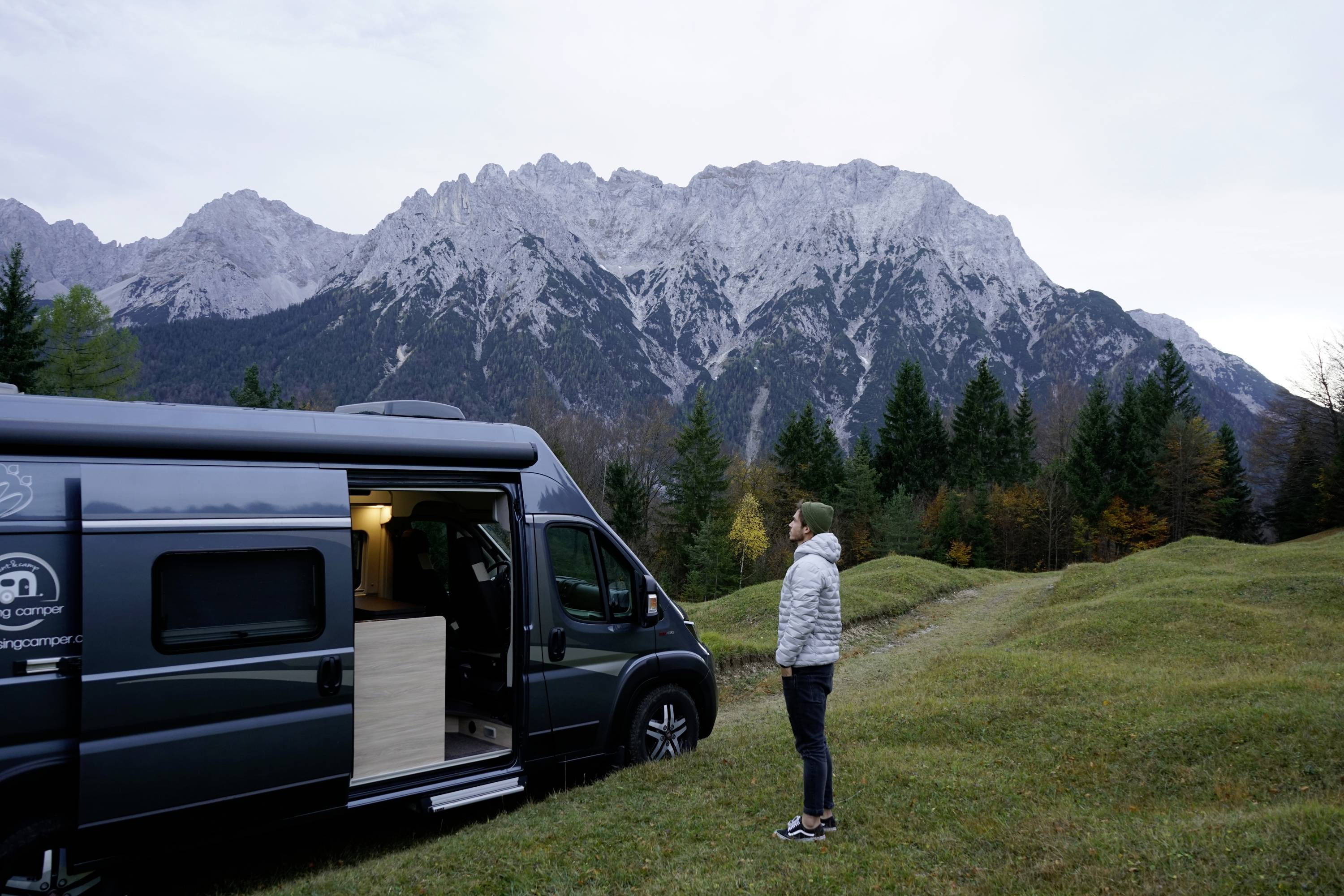 Eine Person steht neben einem geöffneten Wohnmobil, blickt auf bewaldete Hügel und hohe, felsige Berge im Hintergrund.