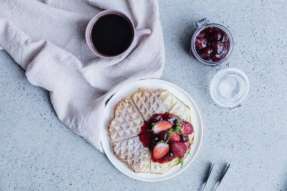 Ein Teller mit Waffeln, bedeckt mit Erdbeeren und Marmelade, daneben eine Tasse Kaffee und ein Glas Marmelade auf einem grauen Tisch.