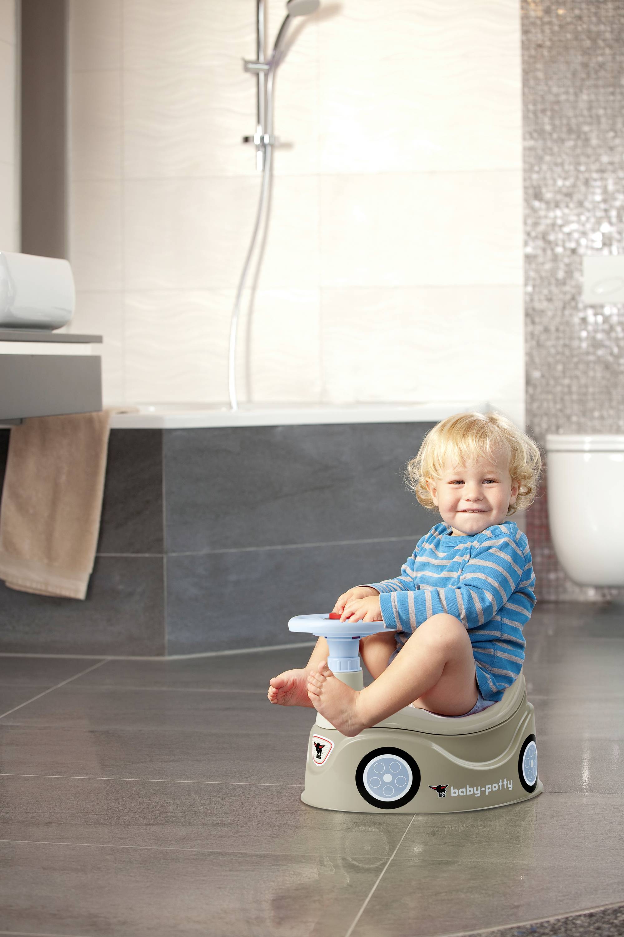 Ein Kleinkind sitzt lächelnd auf einem Töpfchen in einem Badezimmer. Eine Dusche und Toilette sind im Hintergrund zu sehen.