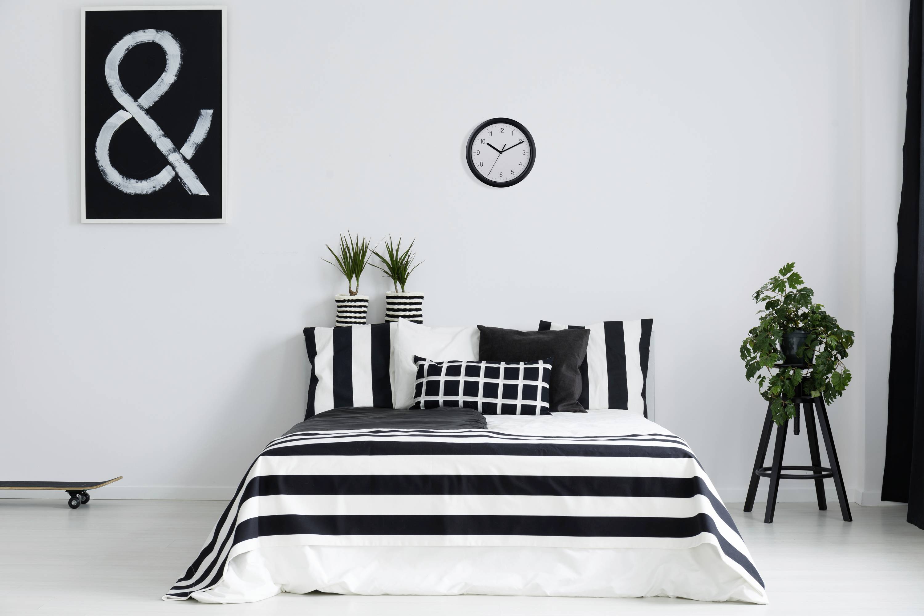 Ein modern eingerichtetes Schlafzimmer mit weiß-schwarzem Farbschema, gestreiftem Bettzeug, zwei Pflanzen auf dem Kopfteil und einer Wanduhr.