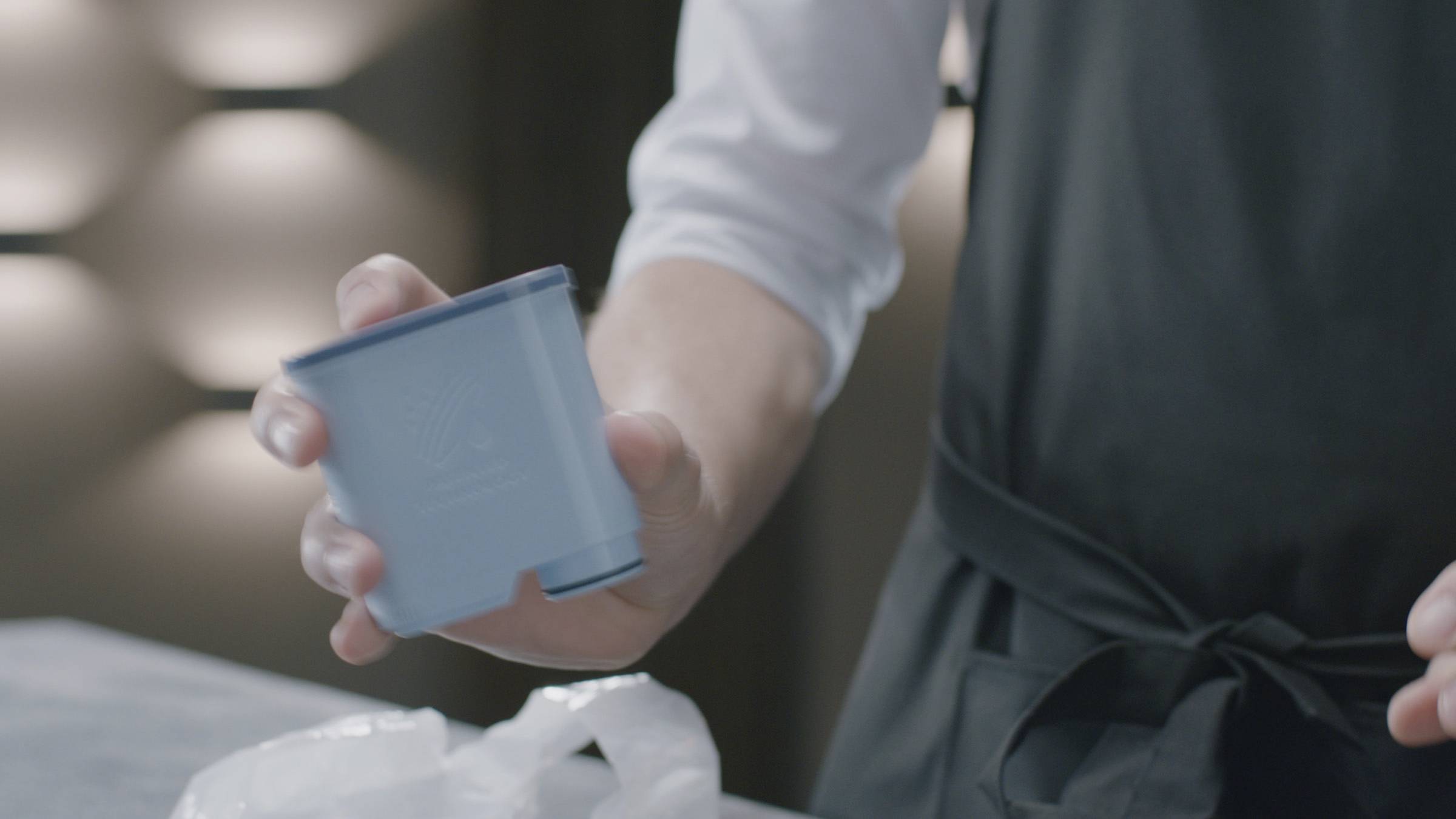 Eine Person hält einen blauen Behälter in der Hand. Sie trägt eine Schürze, und im Hintergrund sind Lichter an einer Wand zu sehen.