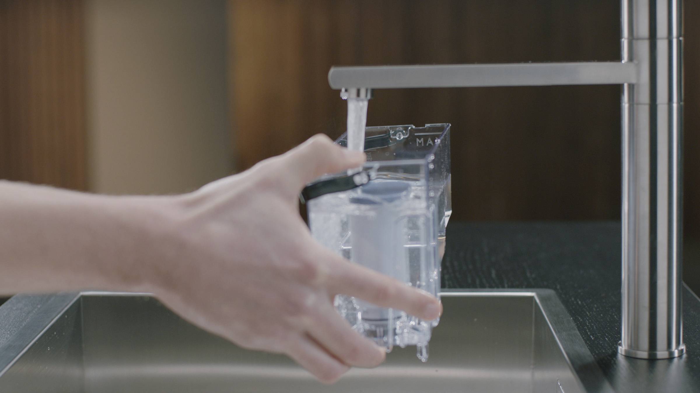 Eine Hand füllt eine durchsichtige Karaffe mit fließendem Wasser unter einem Küchenwasserhahn auf.