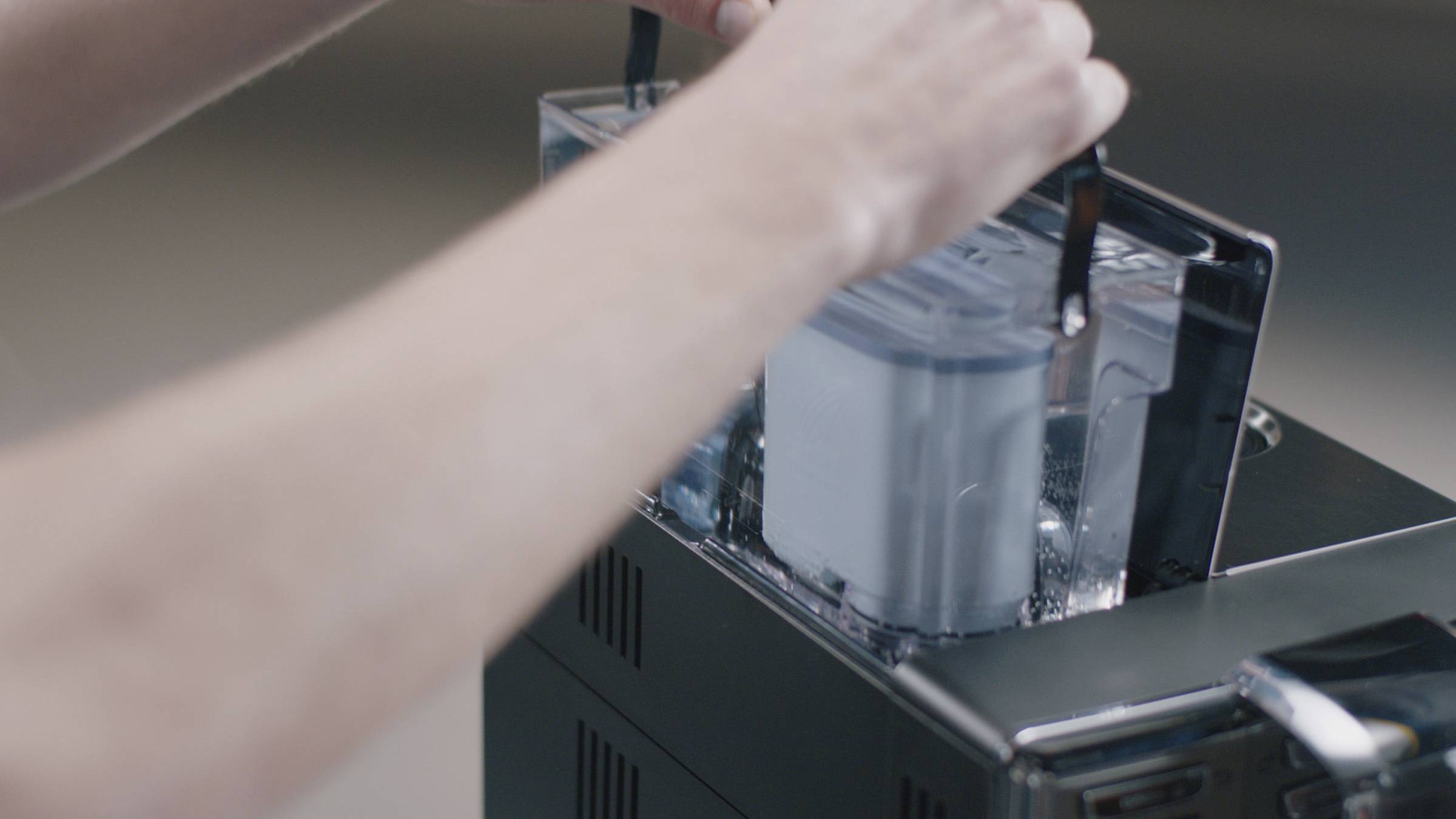 Eine Person setzt einen Wassertank in eine Kaffeemaschine ein. Der Fokus liegt auf den Händen, die den Tank sicher in die Maschine schieben.