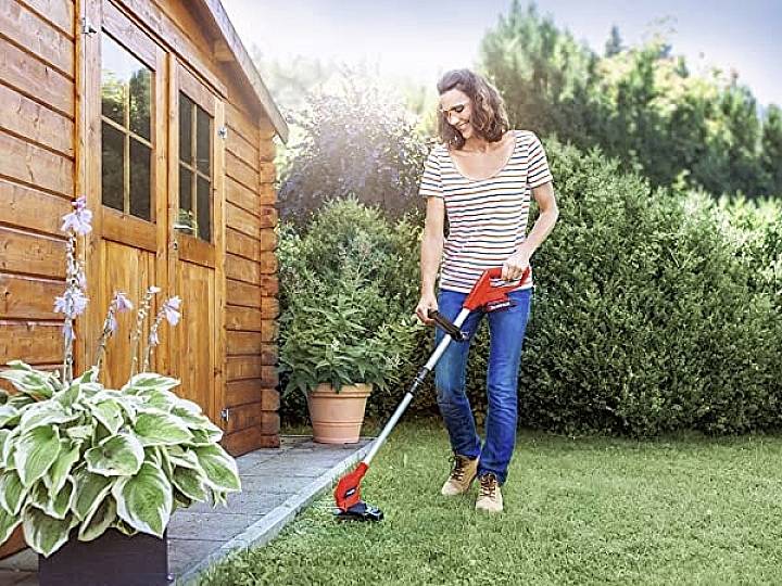 Eine Frau verwendet einen Rasentrimmer, um Rasen in einem Garten neben einem Holzschuppen zu schneiden. Im Hintergrund sind Büsche sichtbar.
