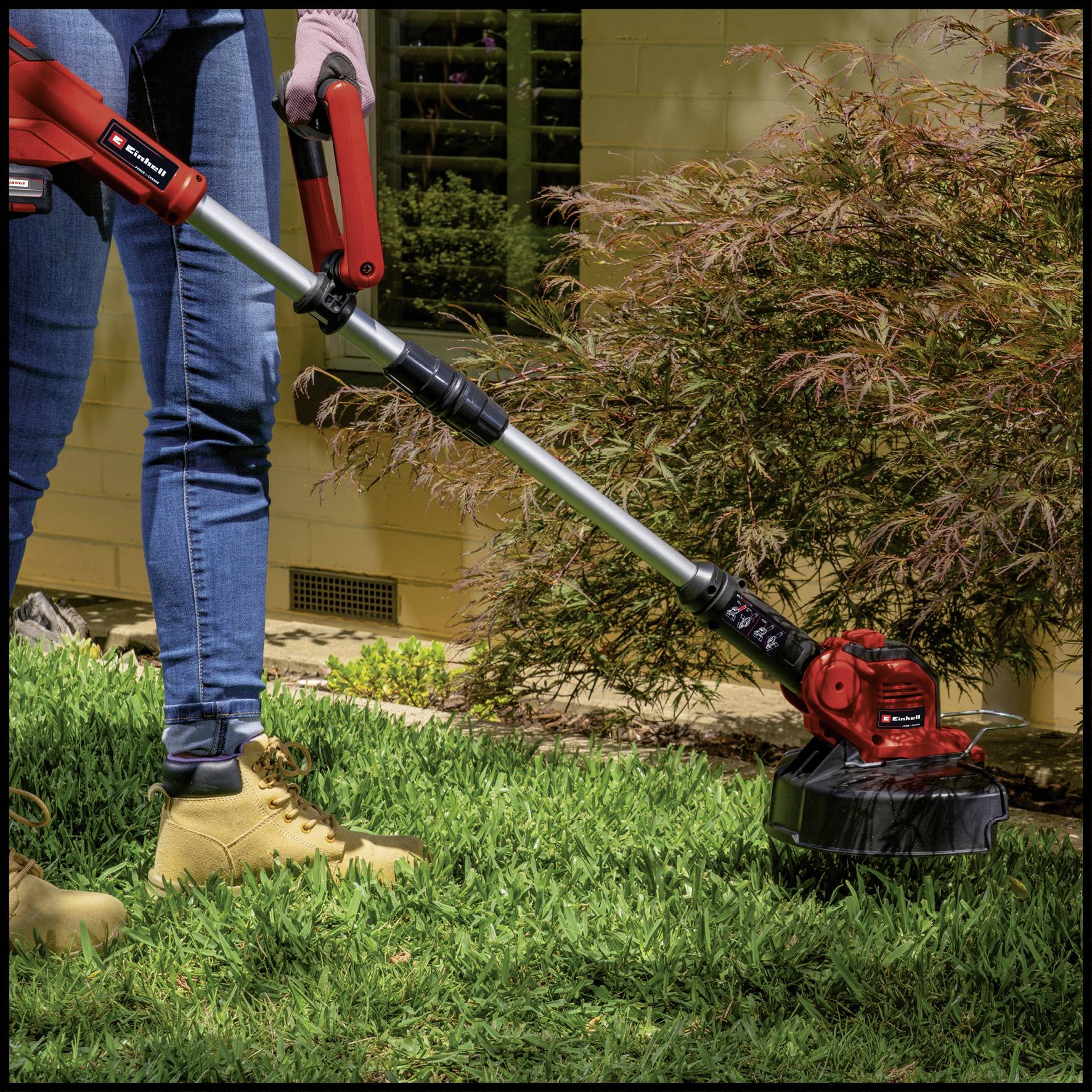 Eine Person in Jeans und Arbeitsstiefeln benutzt einen roten Rasentrimmer, um hohes Gras im Garten zu kürzen.