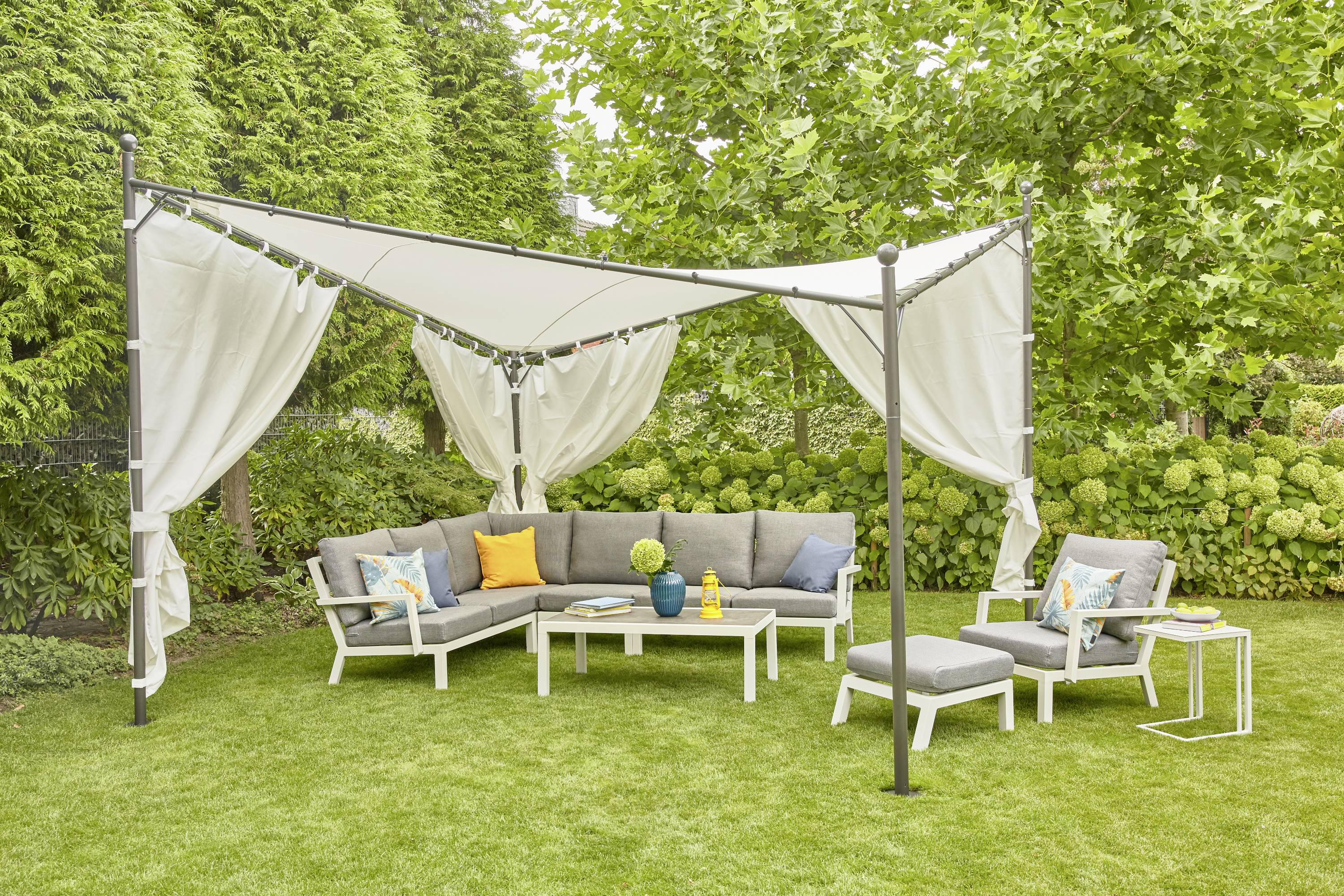 Ein Gartensitzbereich mit grauen Sofas, farbigen Kissen und einem Sonnensegel. Umgeben von grünen Bäumen und Sträuchern.