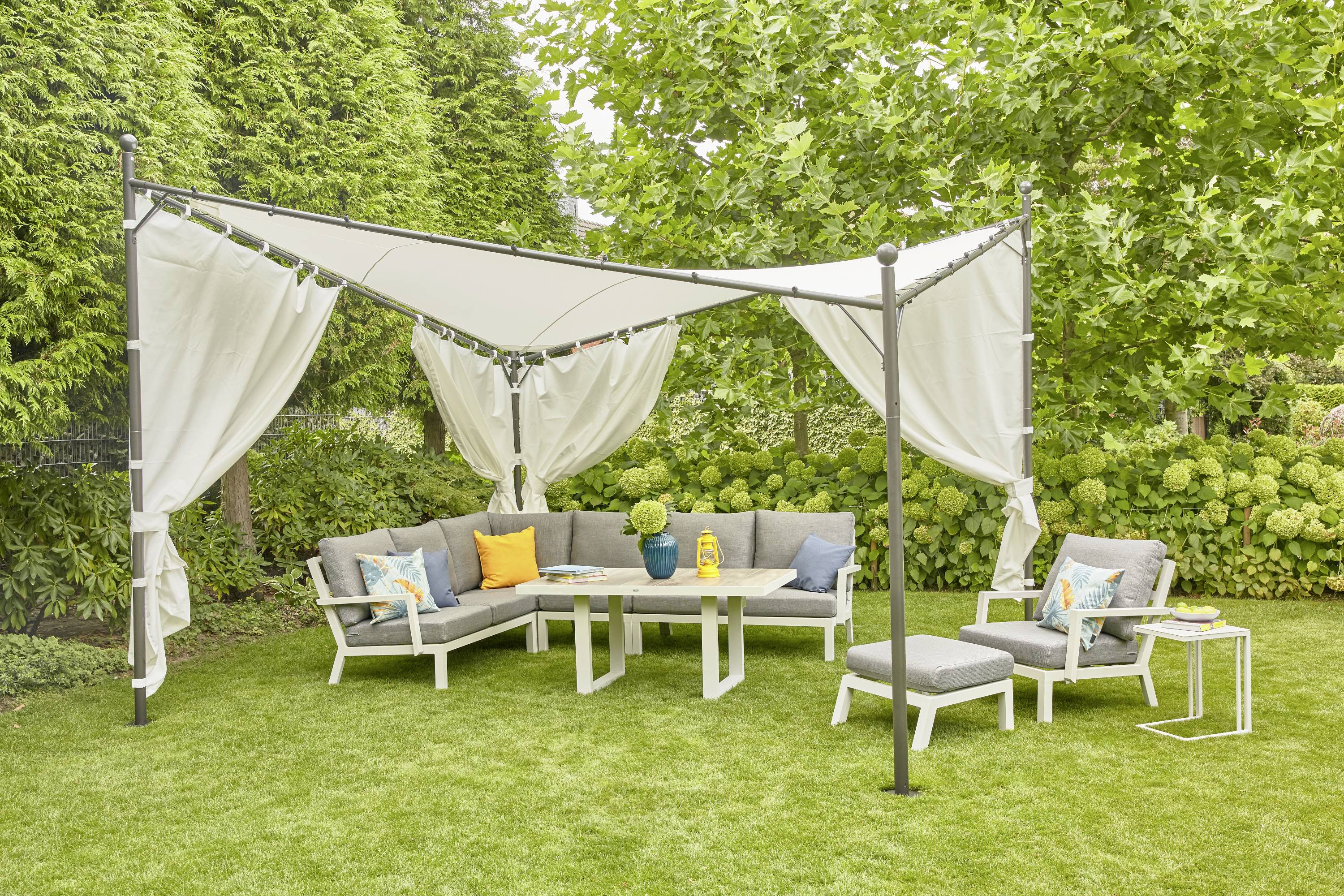 Sitzecke im Garten unter einem Pavillon mit weißen Vorhängen, umgeben von Grün. Graue Sofas und farbige Kissen bieten gemütlichen Sitzplatz.