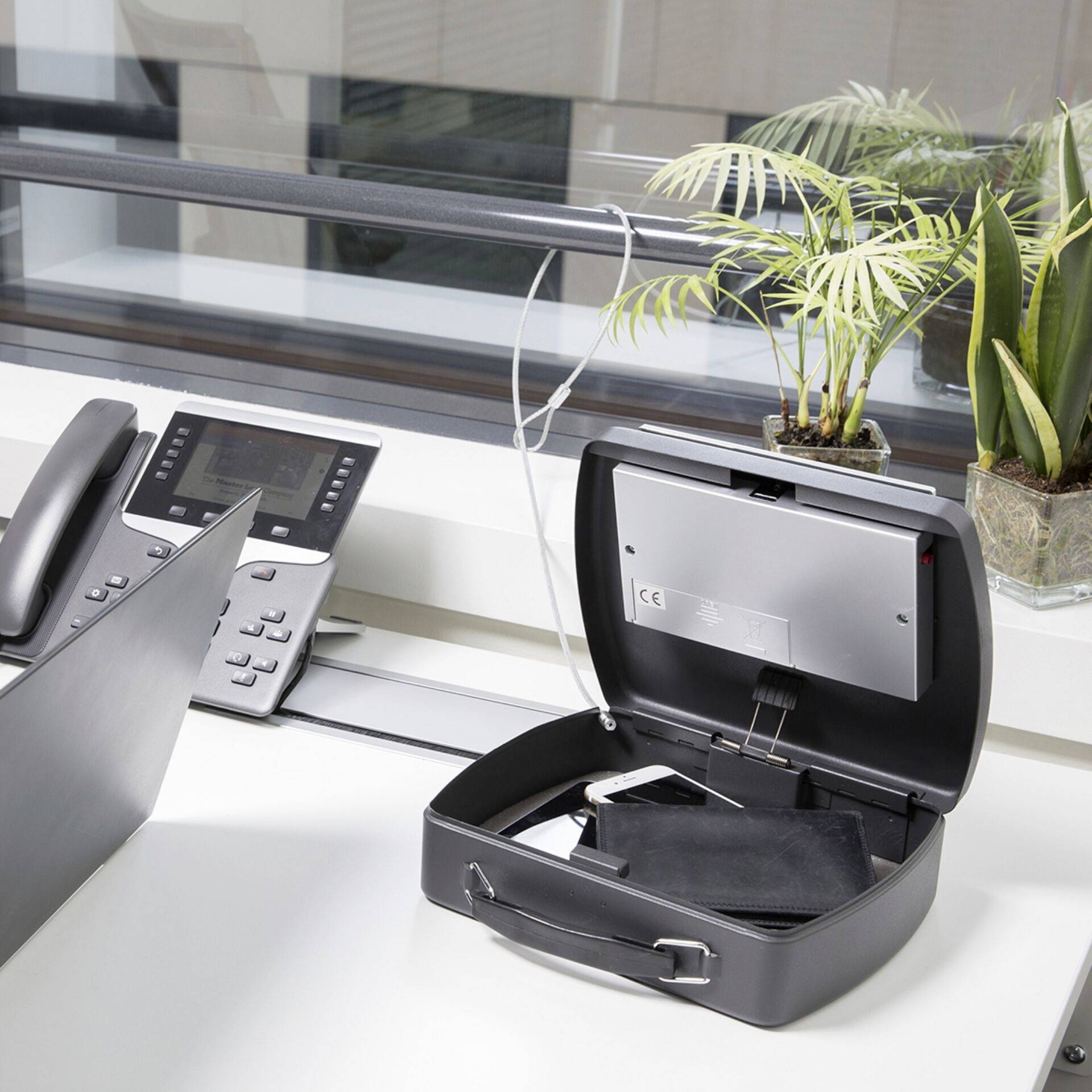 Ein offener Safe auf einem Schreibtisch mit einem Telefon, nahe einem Fenster, daneben eine kleine Pflanze.