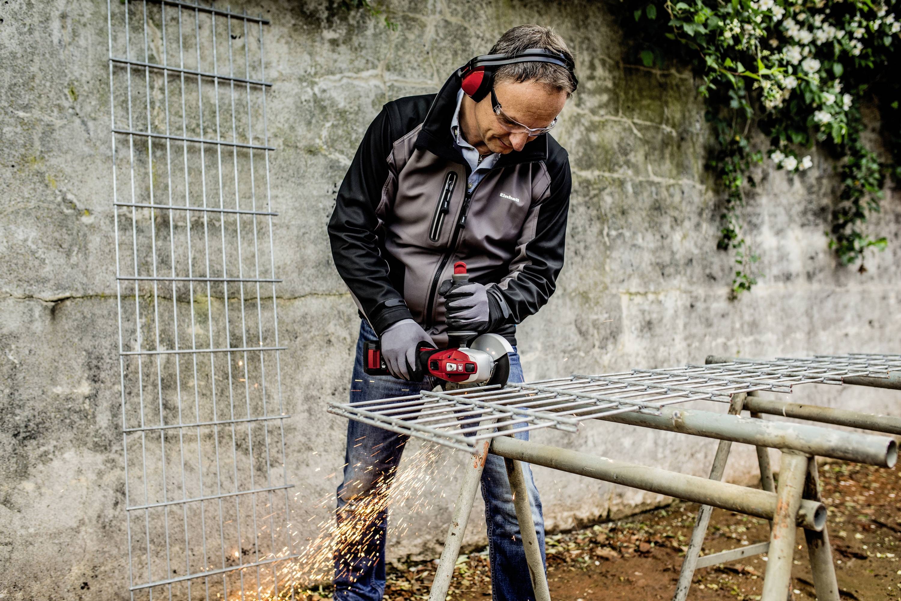 Ein Mann mit Gehörschutz und Schutzbrille schneidet mit einem Winkelschleifer ein großes Metallgitter. Funken fliegen.