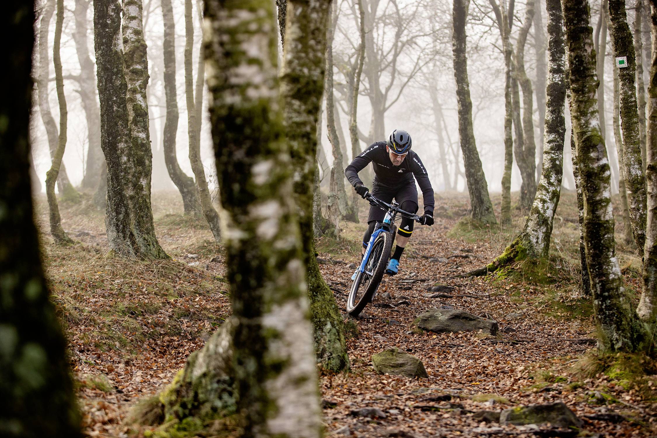 Ein Mountainbiker fährt durch einen nebligen, herbstlichen Wald auf einem schmalen Pfad zwischen Bäumen und über unebene Steine.