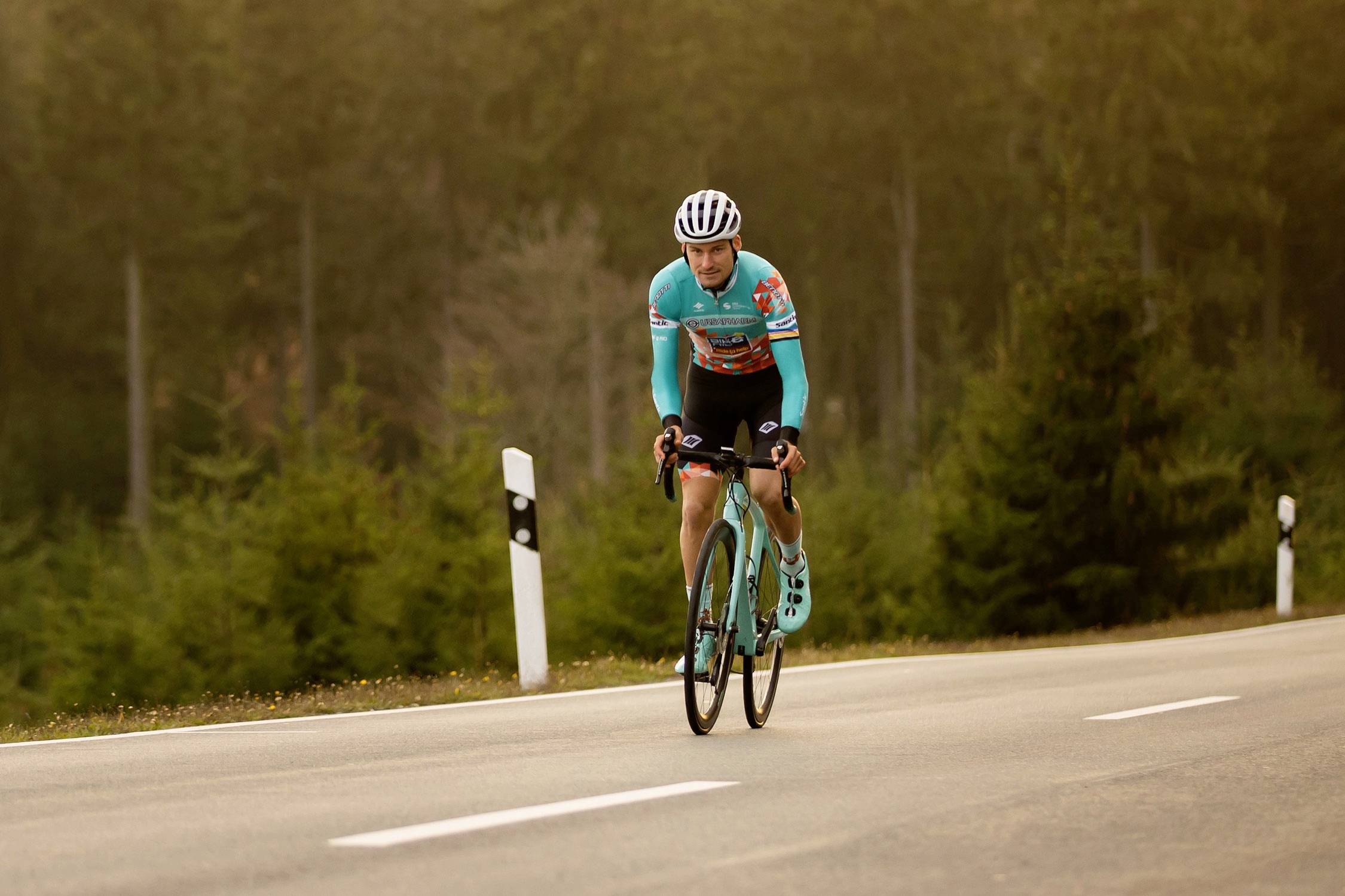 Ein Radfahrer in blau-oranger Sportkleidung fährt auf einer leeren Landstraße durch eine bewaldete Gegend mit Tannenbäumen.