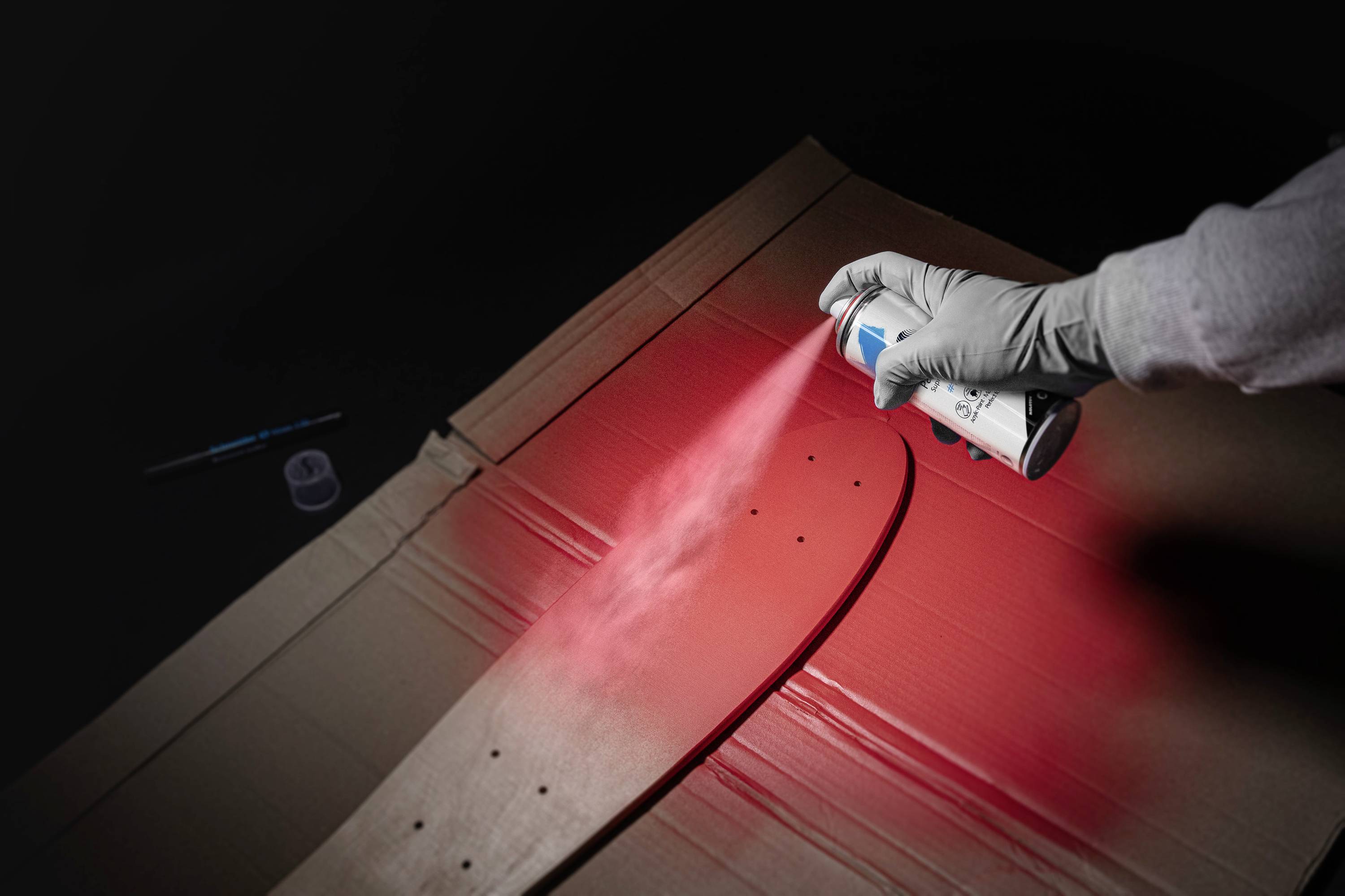 Eine Hand trägt rote Sprühfarbe auf ein Skateboard-Deck auf, das auf einem Karton liegt. Im Hintergrund ein Pinsel und ein Farbtiegel.