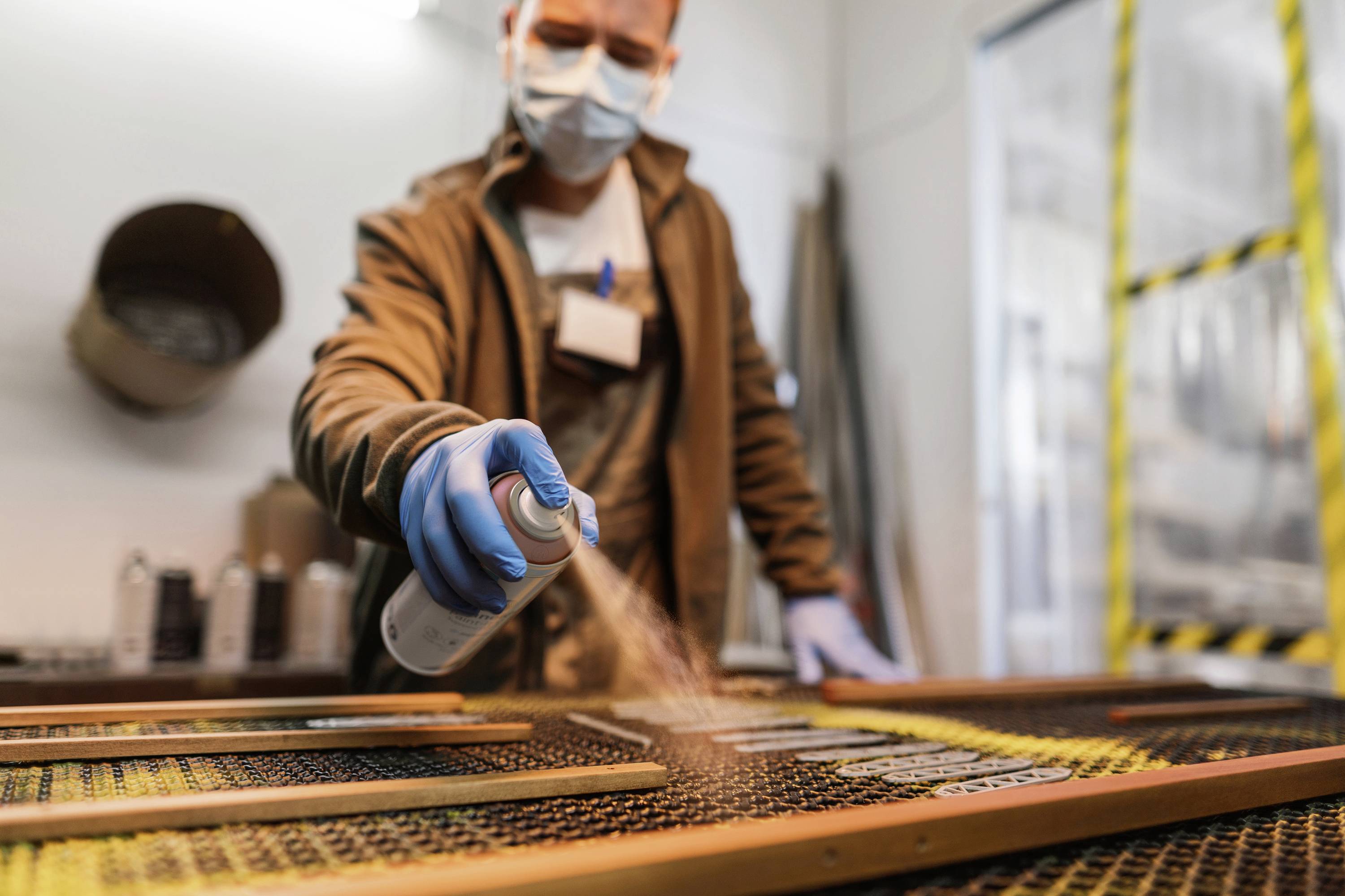 Eine Person in Schutzkleidung sprüht Farbe auf Holzrahmen in einer Werkstatt. Sicherheitsausrüstung und Werkzeuge im Hintergrund.
