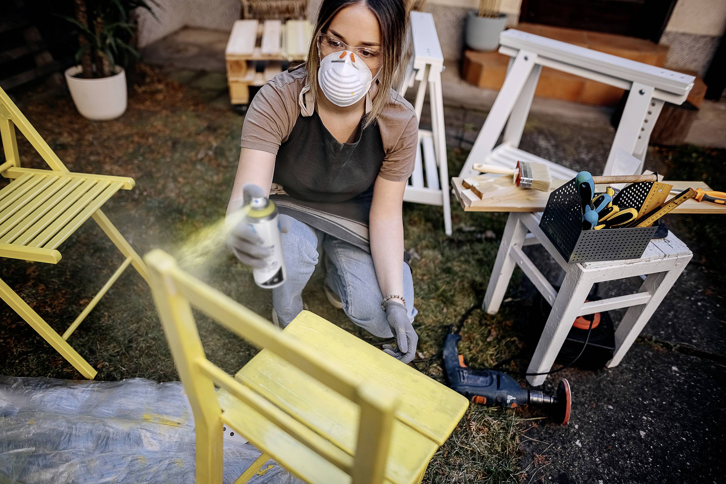 Eine Person sprüht gelbe Farbe auf einen Holzstuhl im Freien. Umgebung: Garten mit weiteren Möbelstücken und Werkzeugen.