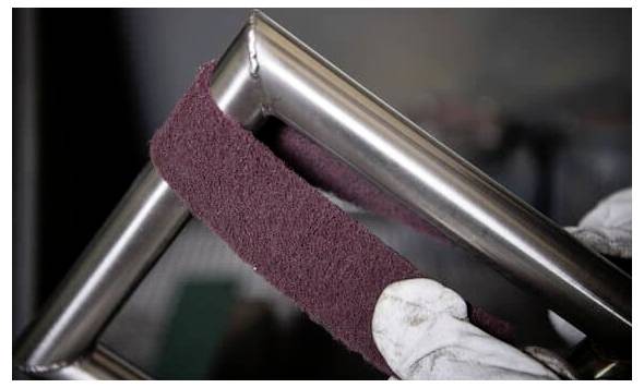 Eine behandschuhte Hand poliert eine Edelstahlrohr-Ecke mit einem burgunderroten Schleifpad und zeigt Metal-Finish- oder Wartungsarbeiten.