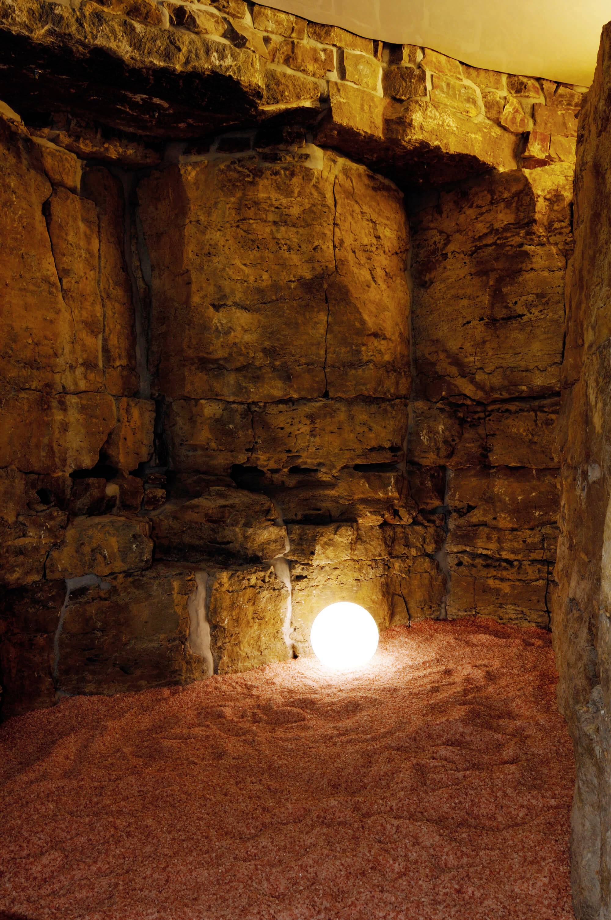 Natürliche Grotte mit erleuchteter Kugel auf dem Boden, umgeben von steinernen Wänden. Atmosphärisches Licht schafft eine warme Stimmung.