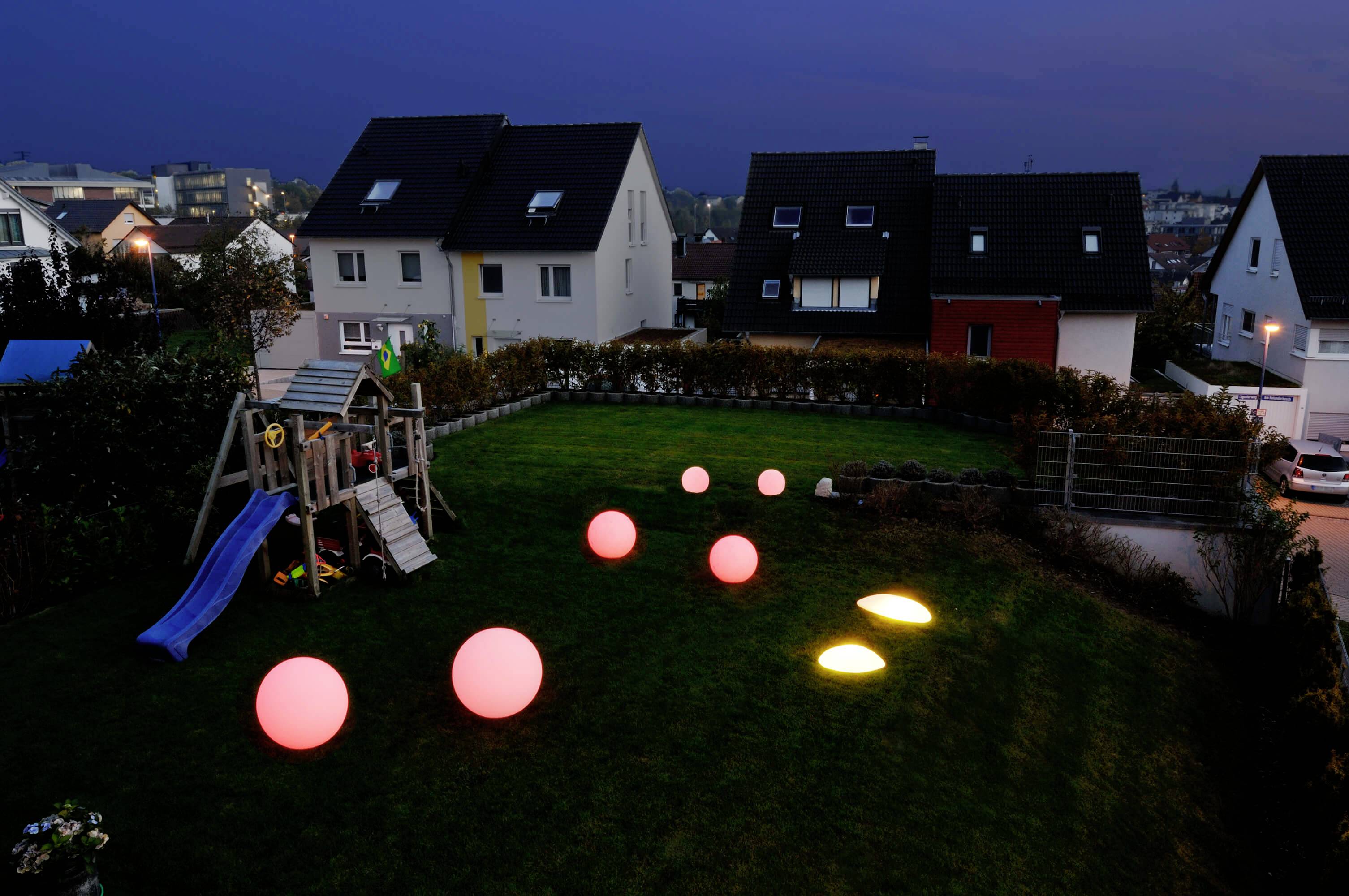 Ein Garten bei Nacht mit Spielplatz und mehreren leuchtenden Kugeln auf dem Rasen, umgeben von Häusern im Hintergrund.
