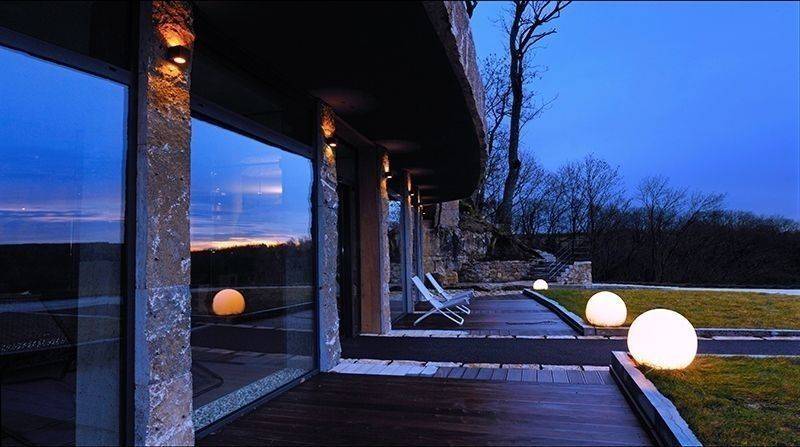 Landschaftsansicht mit modernem Gebäude im Vordergrund, großen Fenstern und beleuchteten Kugeln entlang einer Holzterrasse bei Abenddämmerung.