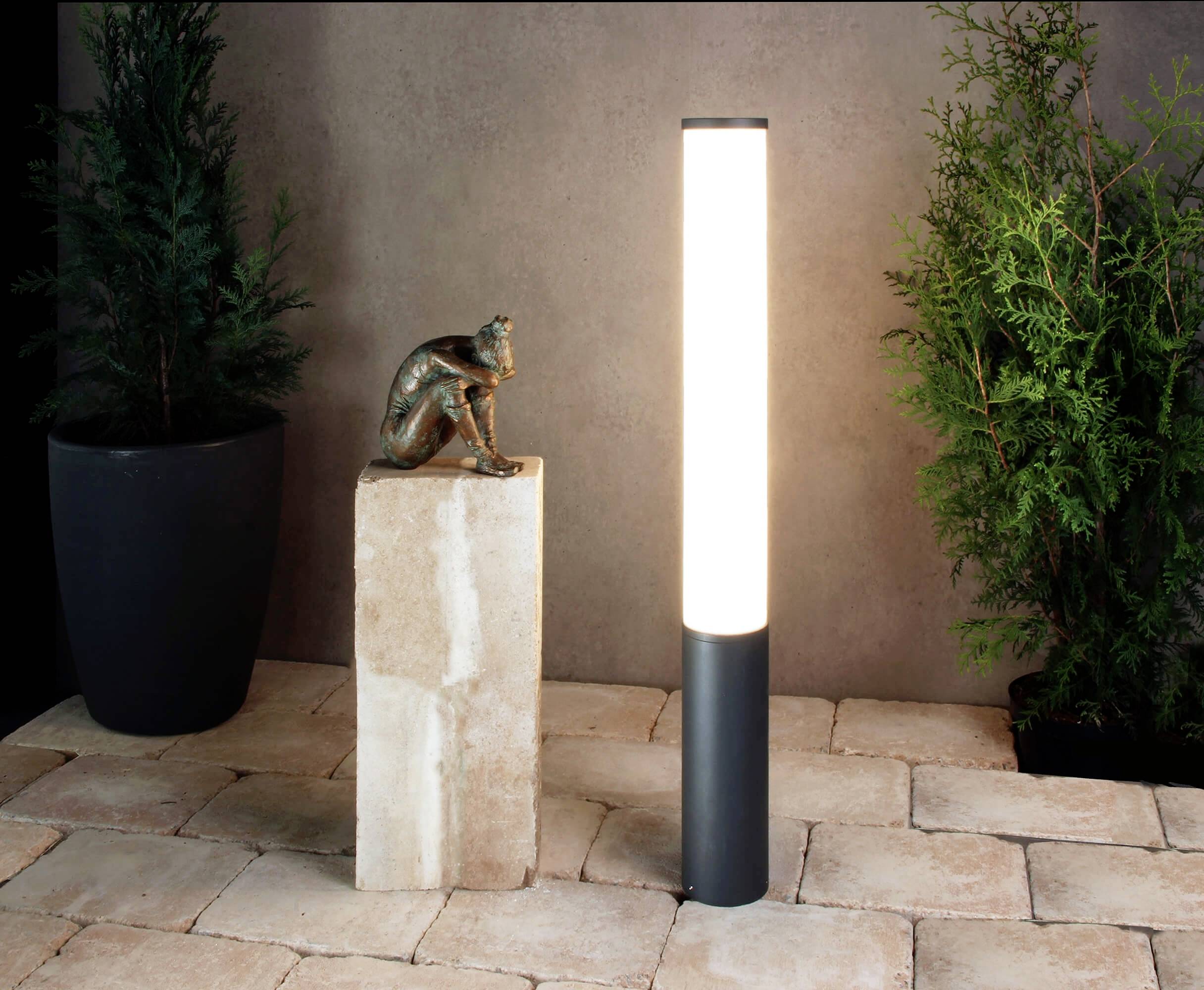 Eine leuchtende Stehleuchte neben einer kleinen Statuette auf einem Steinblock, umgeben von Pflanzentöpfen auf Fliesenboden.