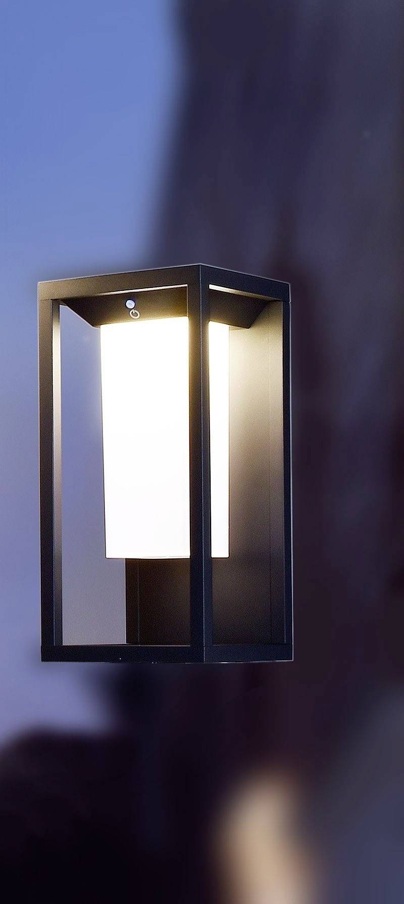 Moderne Wandleuchte mit rechteckigem Rahmen und hellem, diffusem Licht. Sie hängt an einer glatten Wand in der Abenddämmerung.