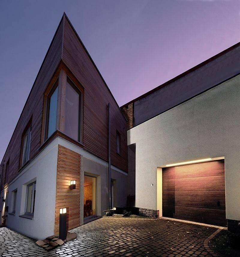 Ein modernes, spitzwinkliges Gebäude mit Holz- und Putzfassade, beleuchtet bei Dämmerung, befindet sich neben einer gepflasterten Auffahrt.