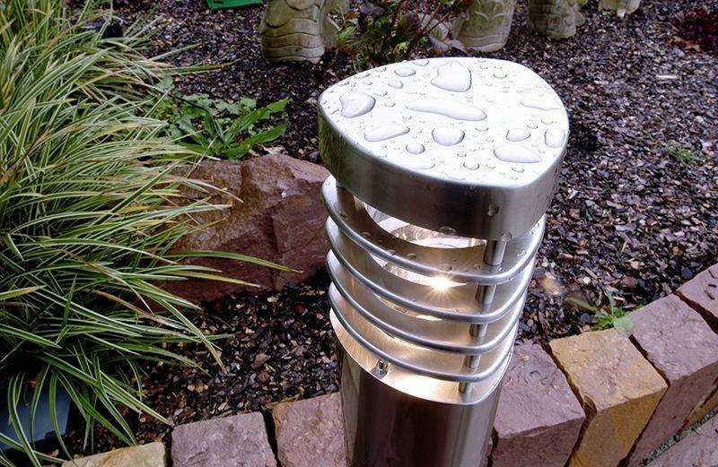 Gartenleuchte aus Metall mit Wassertropfen bedeckt, umgeben von Pflanzen und Steinen.