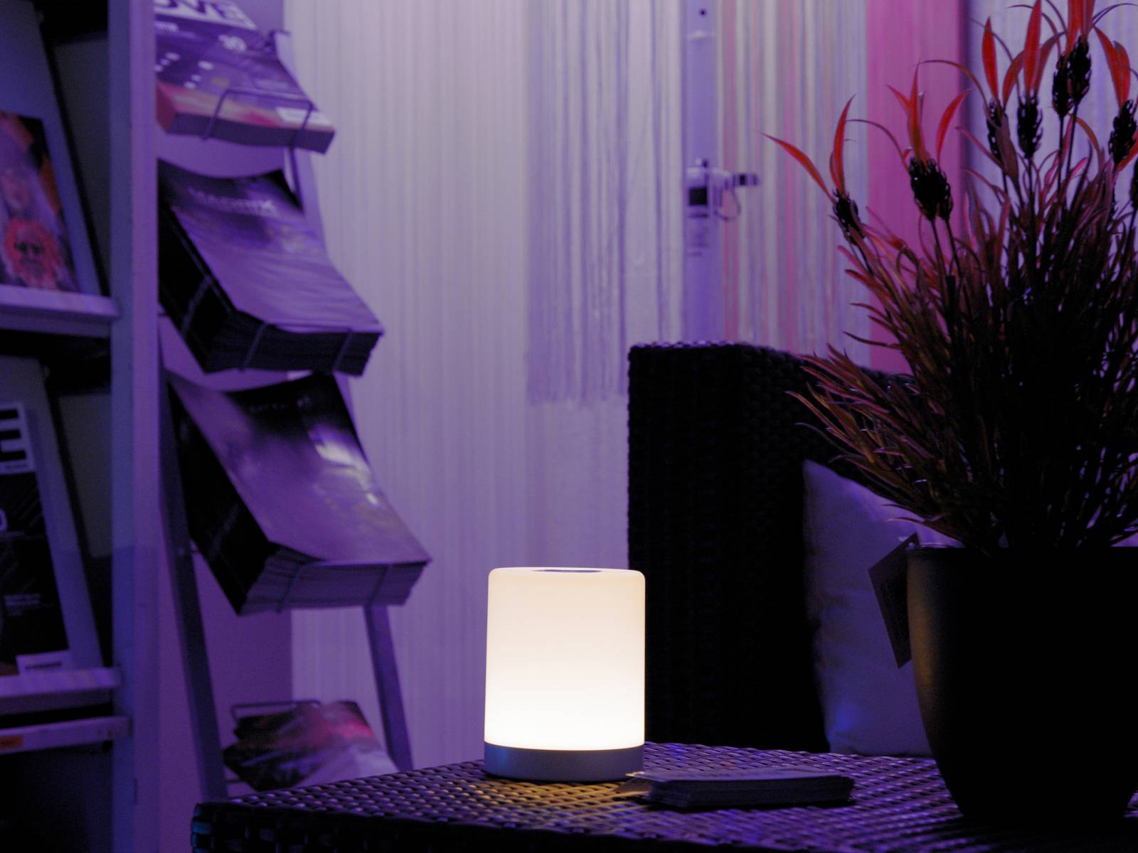 Eine leuchtende Tischlampe auf einem dunklen Tisch in einem Raum mit Zeitschriftenständer und Pflanze im Hintergrund.