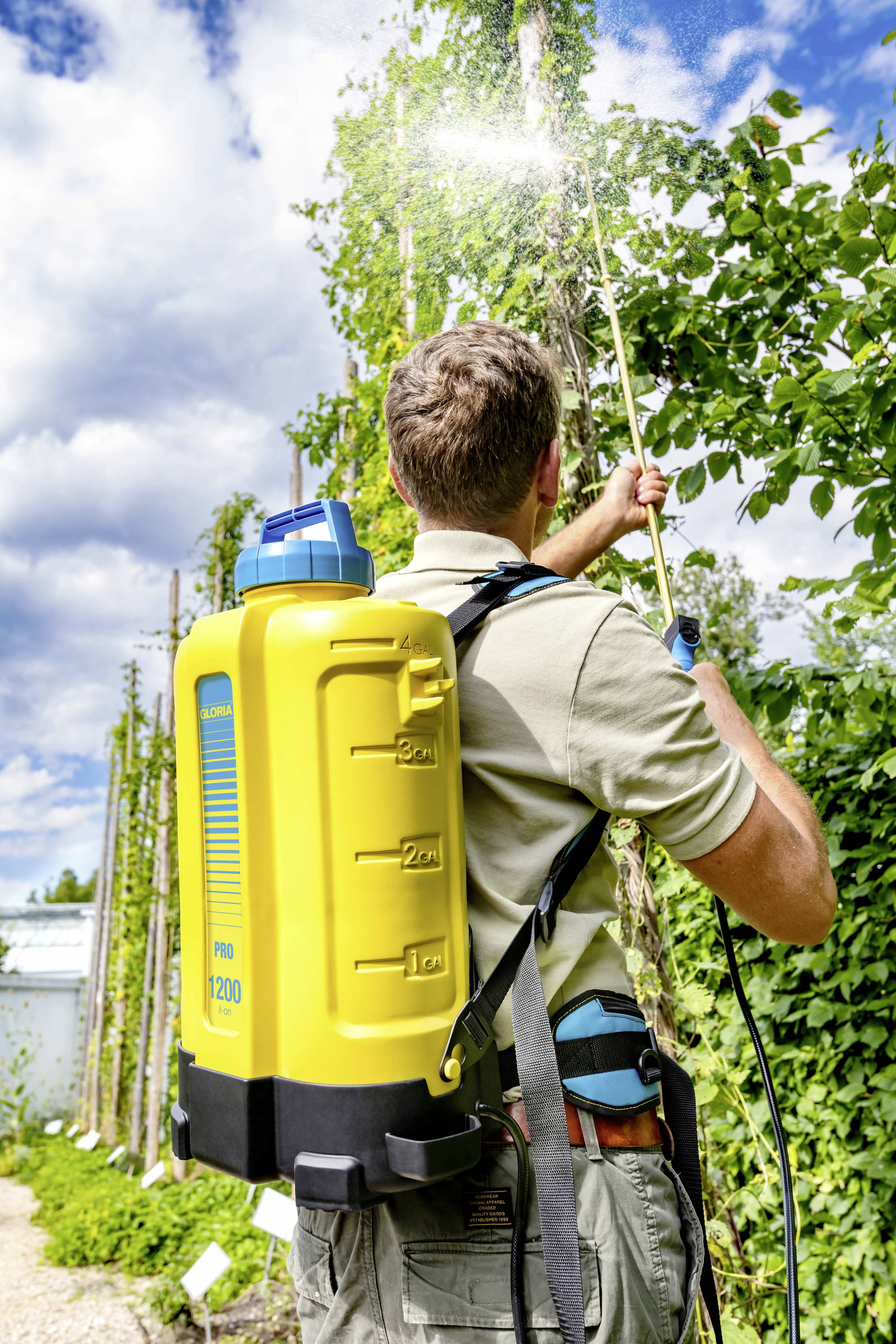 Person sprüht mit einem gelben Rückensprühgerät auf hohe Pflanzen in einem Garten. Der Himmel ist leicht bewölkt.