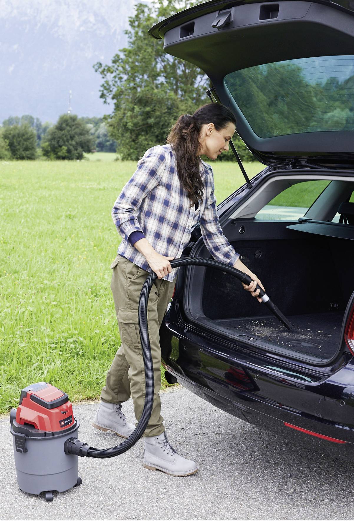 Eine Frau saugt mit einem Staubsauger den Kofferraum eines Autos aus. Im Hintergrund ist eine grüne Wiese zu sehen.