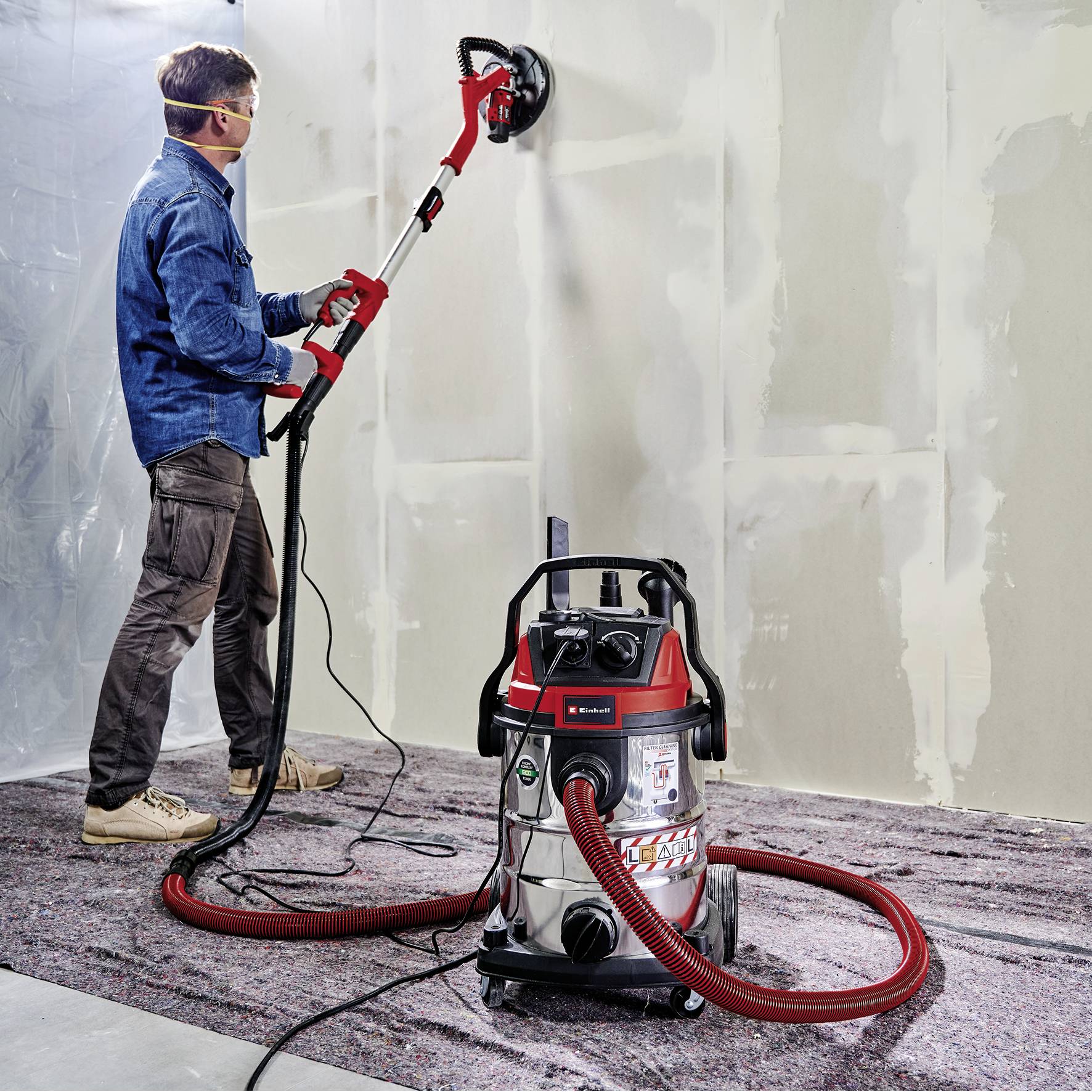 Eine Person schleift mit einem Langhalsschleifer eine Wand. Ein Staubsauger steht auf dem Boden, um Staub zu sammeln.