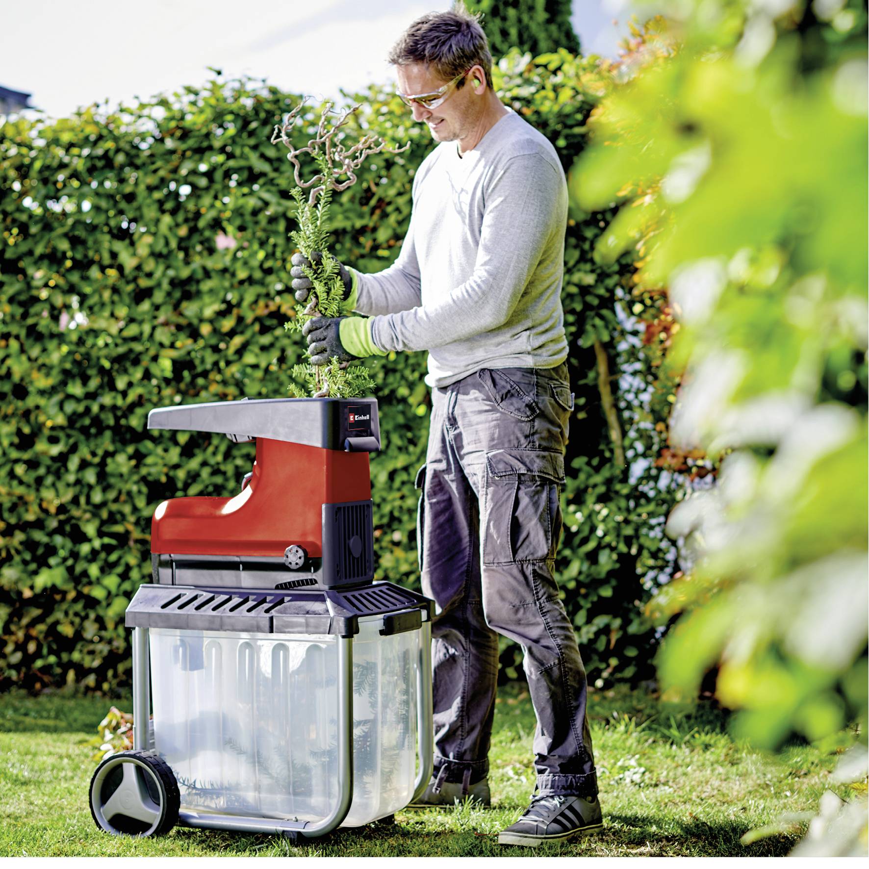 Ein Mann in Arbeitskleidung verwendet einen Gartenhäcksler, um Äste in einem Garten zu zerkleinern. Im Hintergrund sind Hecken zu sehen.