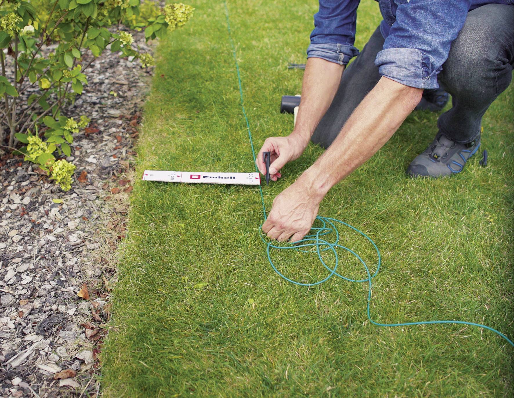 Eine Person legt eine grüne Schnur entlang einer Rasenfläche aus, um eine gerade Linie zu markieren. Ein Maßband und Sträucher sind sichtbar.
