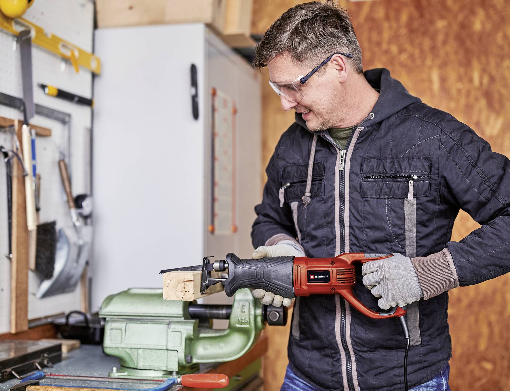 Ein Mann trägt Schutzbrille und Handschuhe, benutzt eine elektrische Säge, um ein Holzstück in einer Werkstatt zu schneiden.