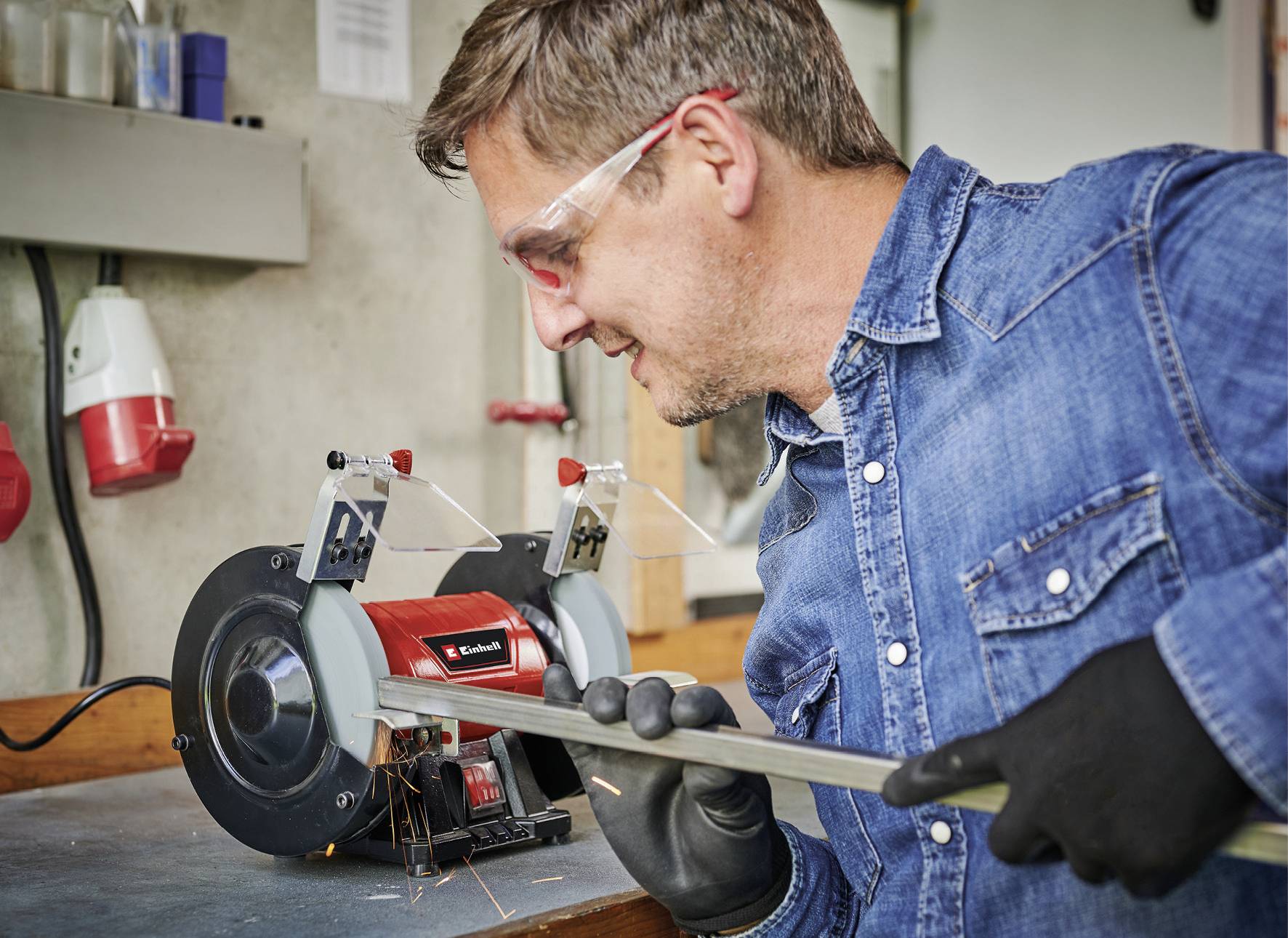 Ein Mann in Schutzbrille und Handschuhen schleift eine Metallstange an einer Werkbank mit einem elektrischen Schleifgerät.