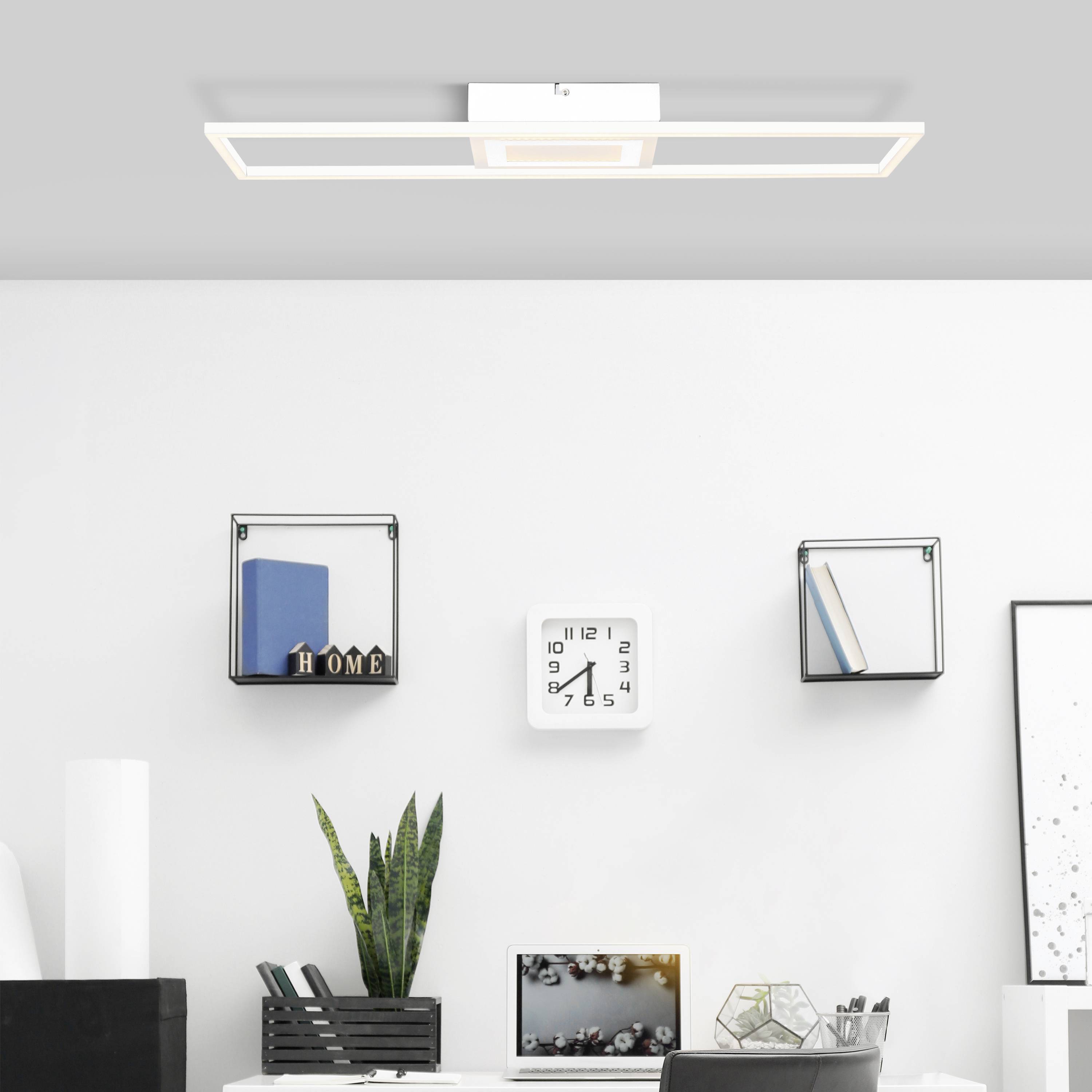 Ein modernes Wohnzimmer mit einer rechteckigen Deckenleuchte, einem Wandregal mit Dekorationen und einem Tisch mit Pflanzen und Büchern.