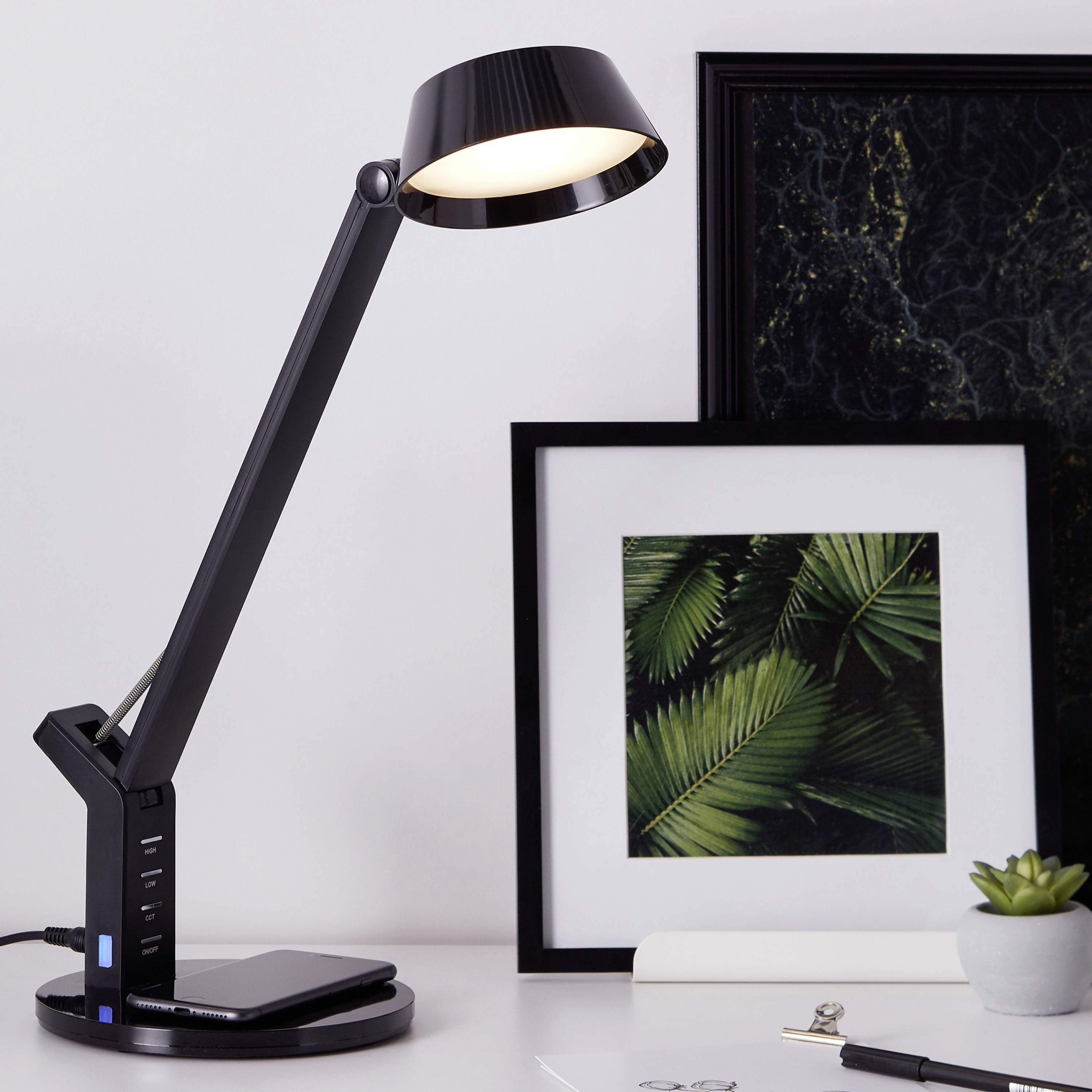 Schreibtischlampe leuchtet auf einen Tisch mit einem gerahmten Foto von grünen Blättern und einem weißen Schreibheft.