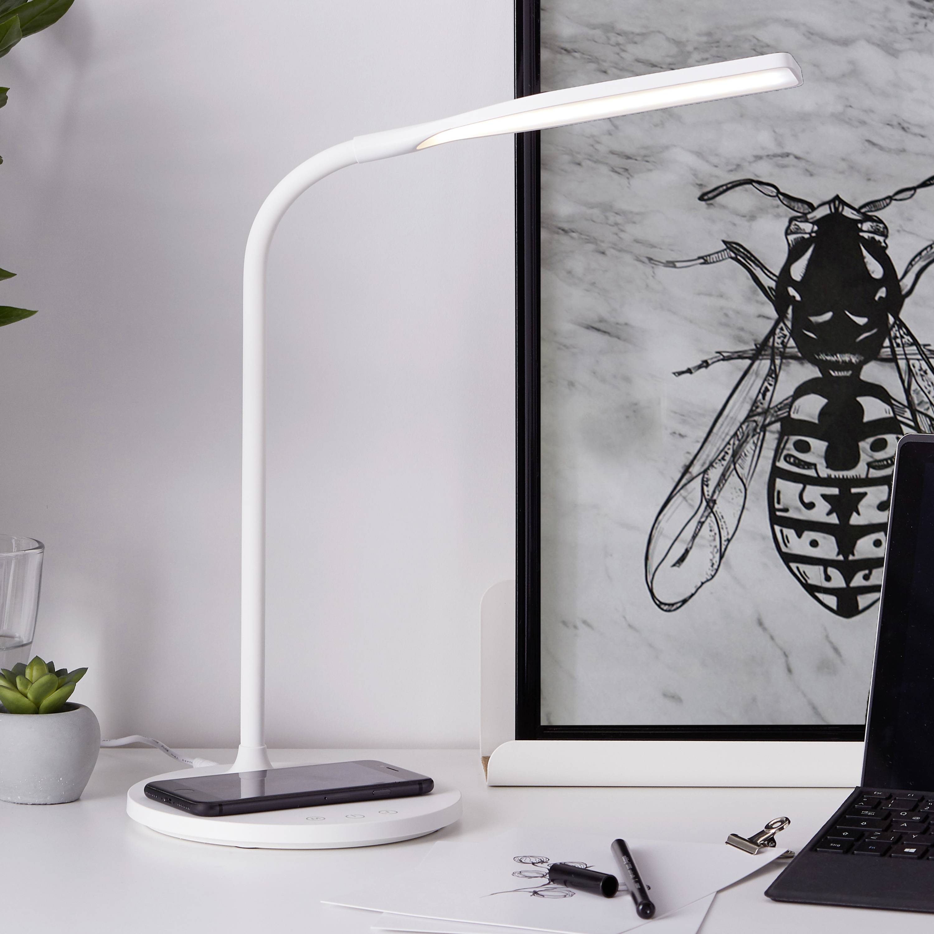Ein stilvoller Schreibtisch mit einer Lampe, die ein Handy drahtlos auflädt. Daneben steht ein Kunstwerk mit einem geometrischen Insektenmotiv im Hintergrund.